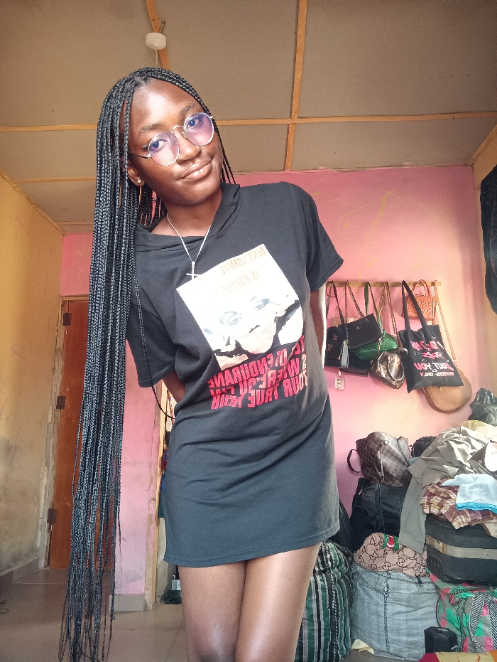 A young woman with long braids and round glasses smiles at the camera. She wears a black t-shirt dress with a graphic on the front. Behind her, a pink wall has purses hanging from a rod. Luggage and other items are piled on the floor. The ceiling is visible, showing a grid pattern.