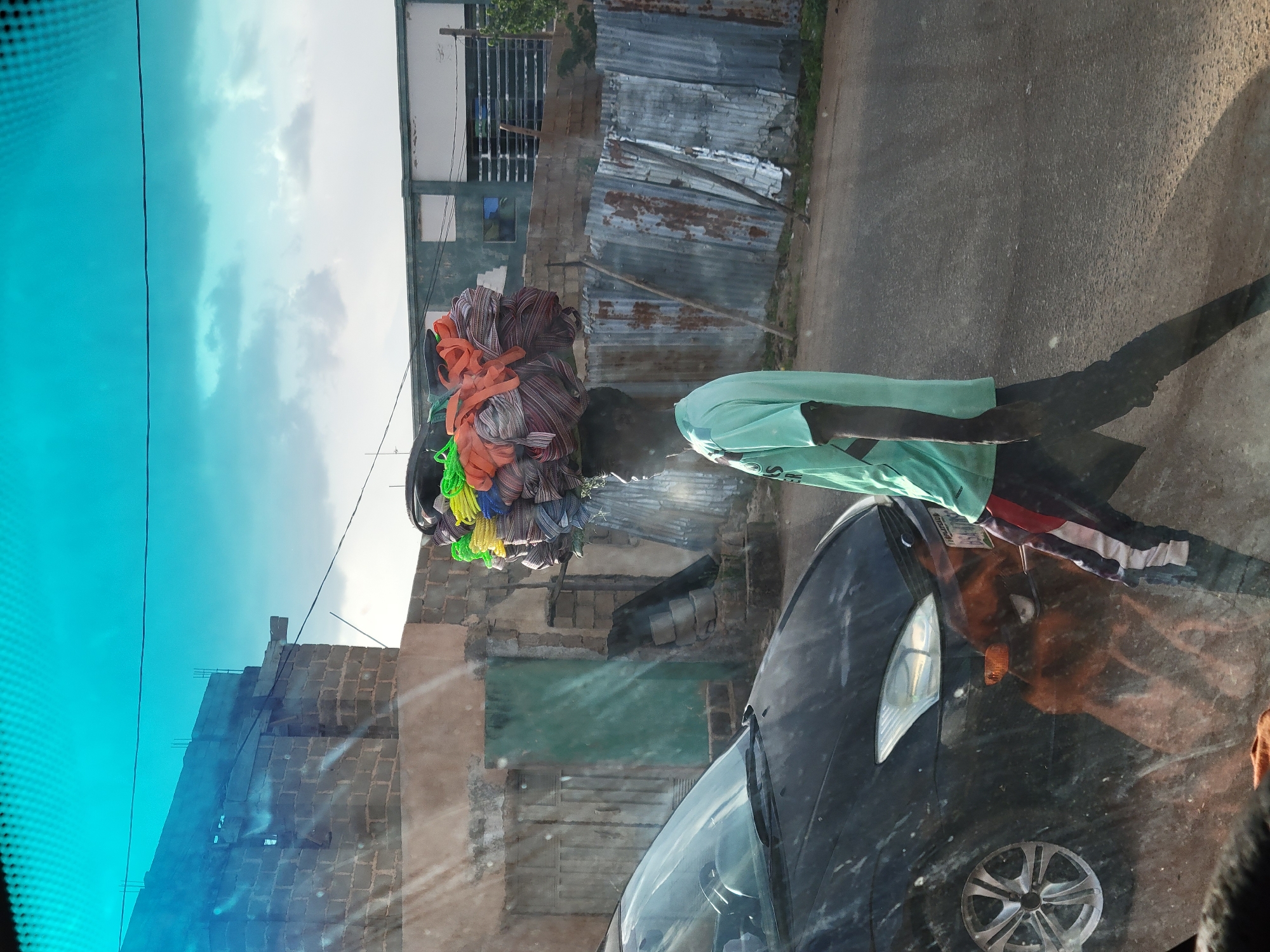 Captured through a vehicle windshield, a street scene in Jos, Nigeria reveals a person carrying a load of colorful materials on their head. The individual, wearing a light green shirt, is walking beside a dark-colored car on an asphalt road. Behind them, a building with a mix of brick and corrugated metal construction is visible. The sky is partially cloudy, with a light blue hue. The vehicle's windshield adds a bluish tint to the overall image.