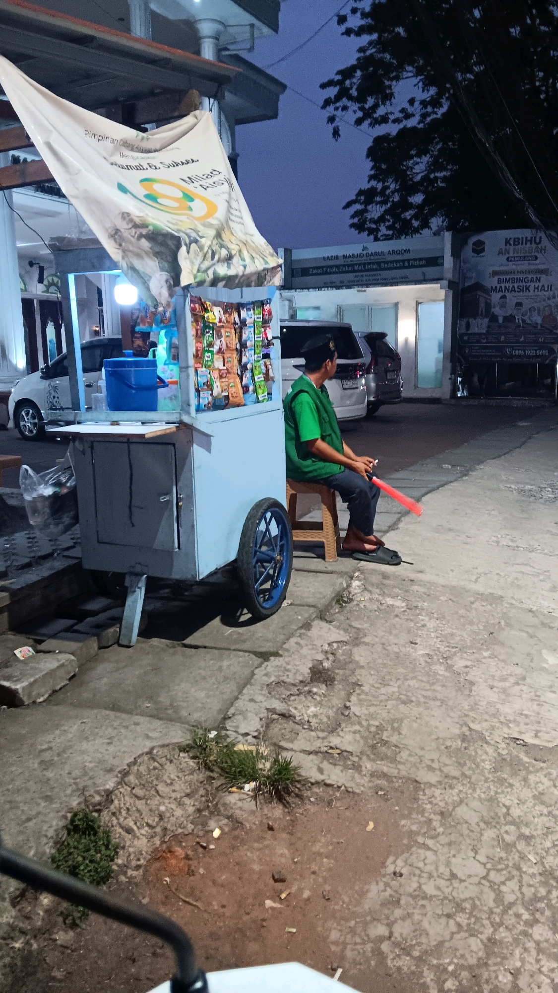 In South Tangerang, Indonesia, a street-side snack cart vendor sits on a brown plastic stool in front of his light blue cart. The cart displays a variety of bagged snacks and drinks. The vendor, wearing a green t-shirt and a black peci cap, is holding a red plastic baton, possibly for directing traffic or for sale. He is barefoot, wearing dark pants and flip-flops. A white banner with Indonesian text and graphics is attached to the front of the cart, partially obscuring the view. In the background, a building with the text "LAZIS MASJID DARUL ARQOM" is visible, along with parked cars and a truck with signage in Indonesian. The scene is lit by ambient streetlights, suggesting it is nighttime or dusk. The ground is paved with concrete, with some patches of dirt and grass.