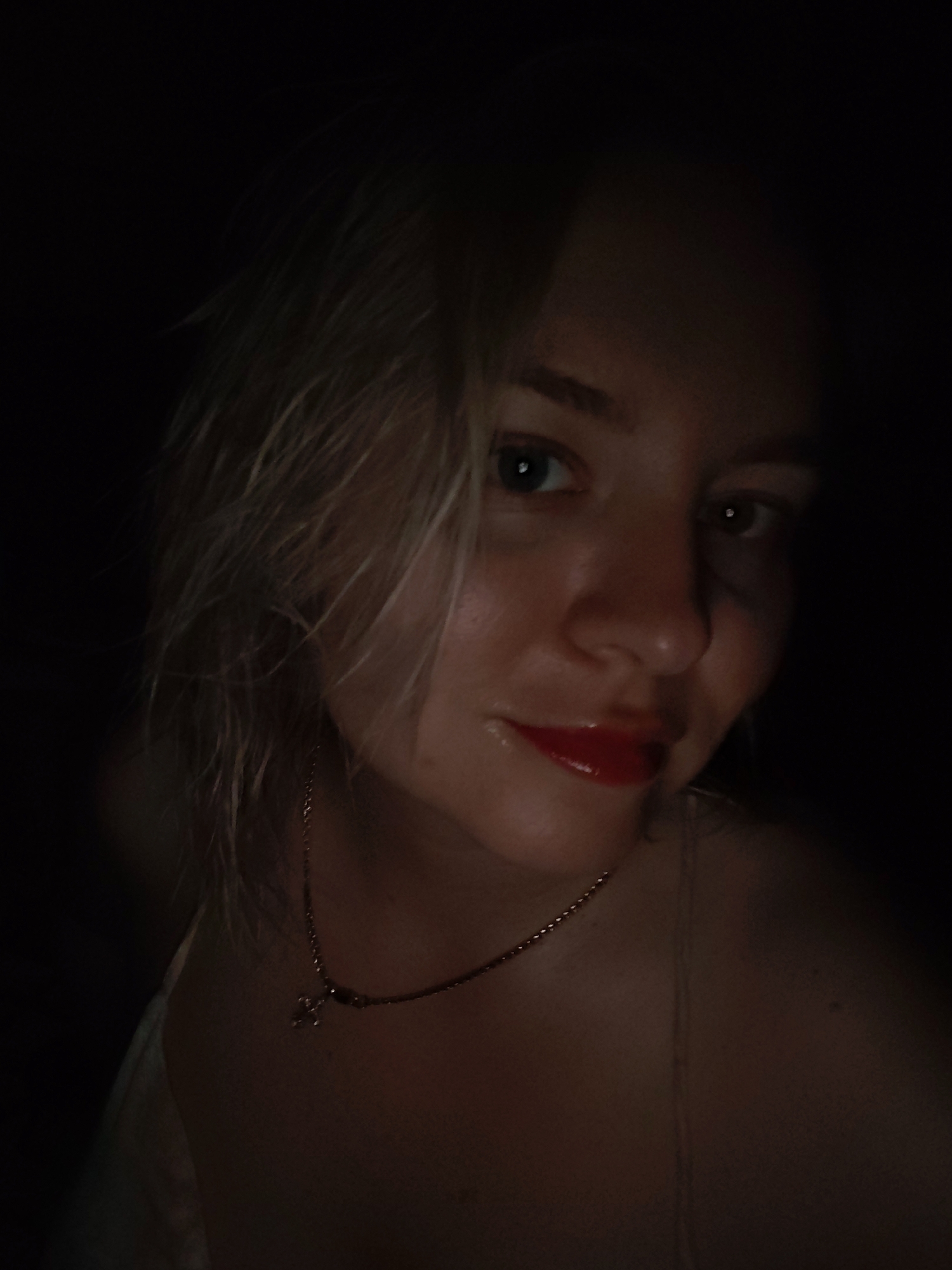A dimly lit, close-up portrait depicts a young, fair-skinned woman with wet, blonde hair. She has light-colored eyes and wears red lipstick. Around her neck, a delicate gold necklace with a small pendant is visible. The background is mostly black, obscuring details. The location in Izhevsk, Russia is not deducible from the image.