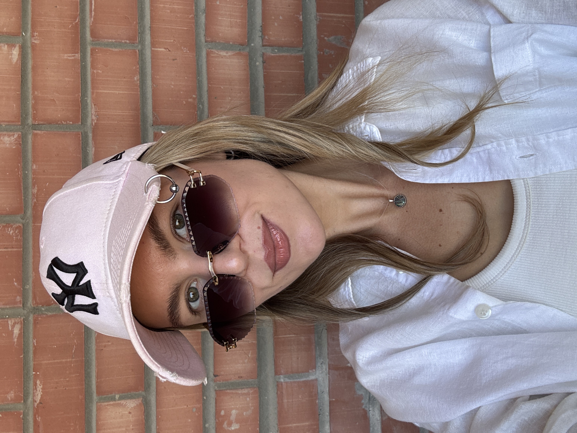 A young Caucasian woman stands against a brick wall, facing the camera directly. She has light brown hair and green eyes, and is wearing a light pink baseball cap with a black logo, oversized sunglasses with dark lenses and gold frames, and a white button-down shirt over a white tank top. She is also wearing a delicate silver necklace with a dark pendant. The woman is smiling slightly. The background is a red brick wall.