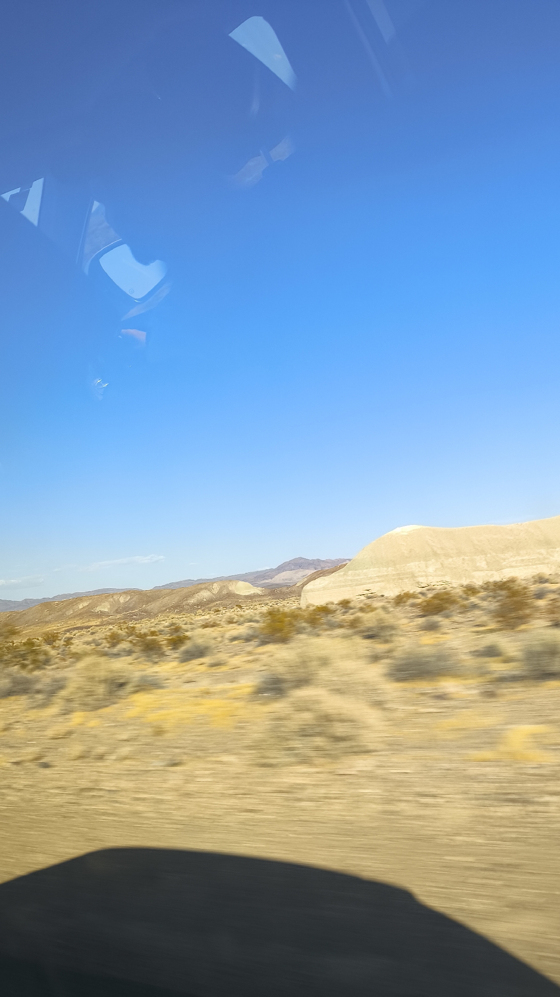 A bright blue sky fills the top portion of the image, reflecting off the car's window. In the distance, desert landscape unfolds with low, rolling hills covered in sparse, dry vegetation. A prominent, pale beige formation, possibly a sand dune or a rock outcrop, dominates the mid-right of the frame. The lower part of the image shows the blurred ground, indicating movement, and a dark shadow, likely from the vehicle itself, stretches across the foreground. The scene appears to be a scenic drive through a desert region, possibly near Tecopa, California, United States, given the characteristic arid terrain.