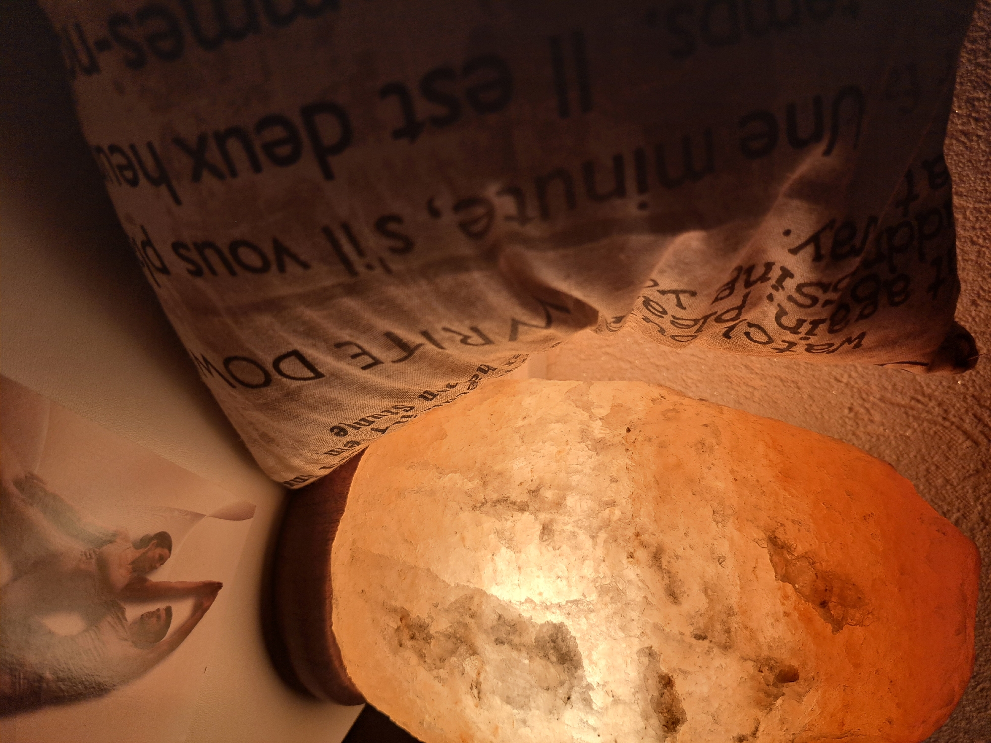 A close-up, dimly lit, indoor scene features a large, glowing Himalayan salt lamp and a textured fabric object with text printed on it. The salt lamp, positioned on the right, is a rough-hewn, translucent orange and white rock emitting a warm, soft light. The fabric object, on the left, is a light-colored material with dark text arranged vertically, partially obscuring it. The text is in French and appears to be a quote. The background is a textured, light-colored wall. In the foreground, a translucent image of two people in a dance pose is visible. The overall impression is one of a peaceful, possibly meditative, atmosphere.