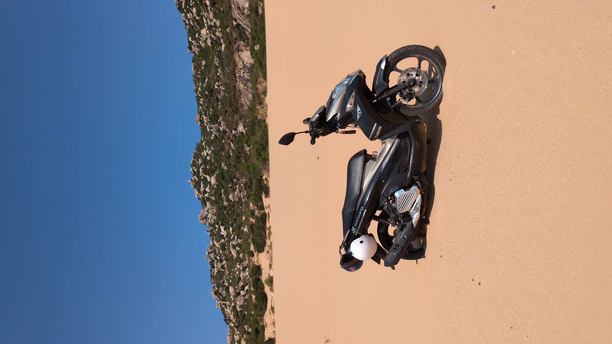 The image shows a Yamaha motorcycle parked in a desert-like landscape in Phuoc Dinh, Vietnam. The bike, a black model, is parked on a sandy surface. A helmet hangs from the side. In the background, a rocky hill dotted with vegetation is visible against a clear blue sky.