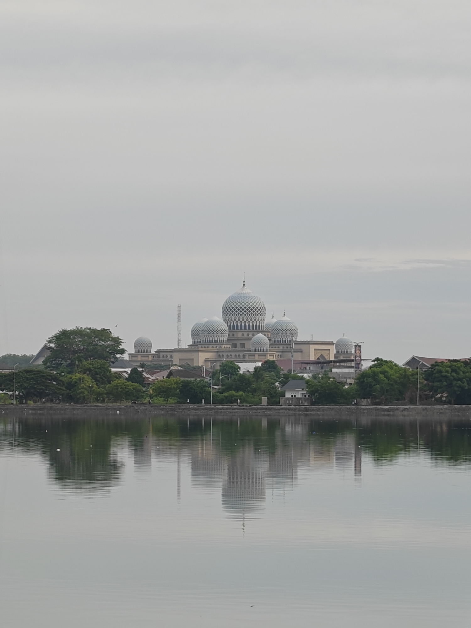 The image depicts a mosque with multiple domes, likely the Islamic Center of Lhokseumawe, Indonesia, situated across a body of water. The mosque's architecture features prominent white domes with intricate patterns. The building is surrounded by trees and other structures, and its reflection is visible on the calm surface of the water. The sky is overcast, suggesting a cloudy day. A few people are discernible walking along the waterfront.