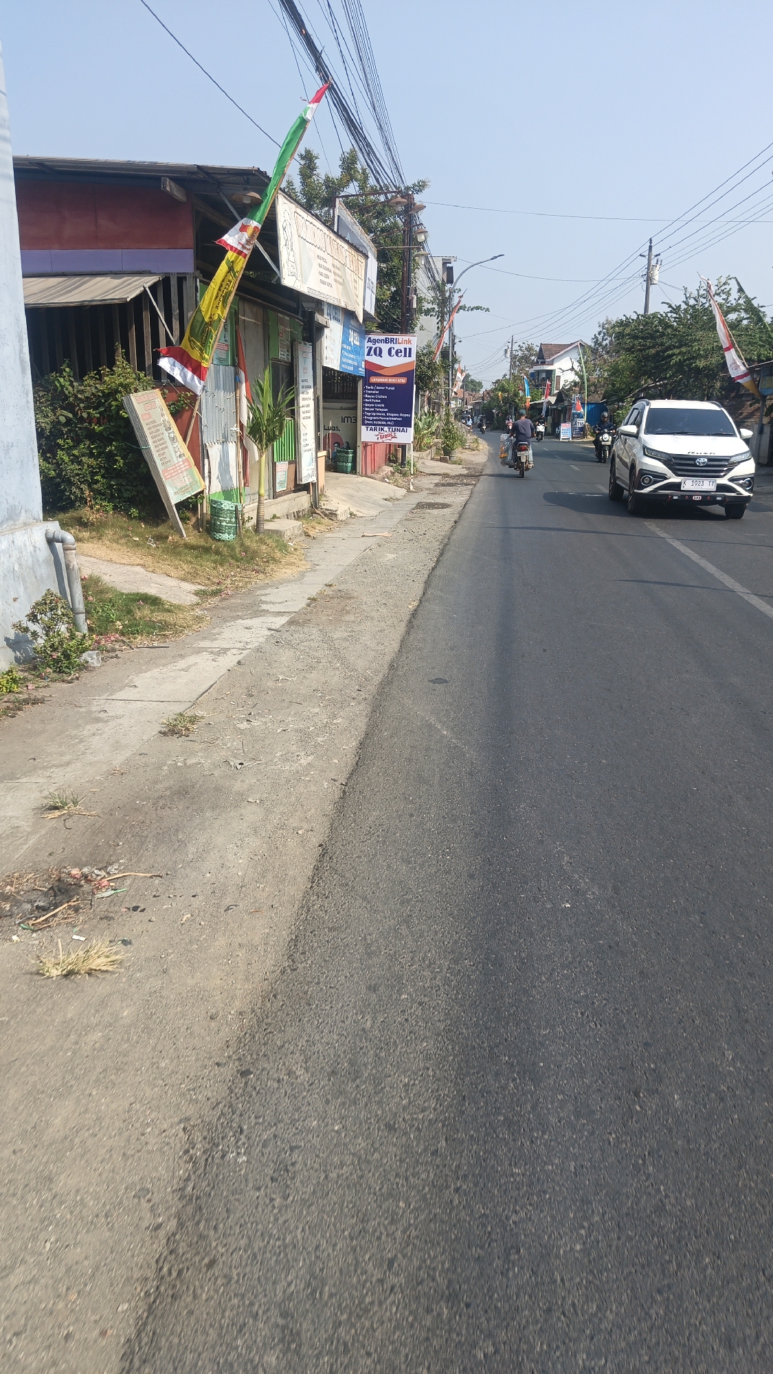 The image depicts a street scene in Bulungcangkring, Indonesia, on a sunny day. On the left side of the road, a building with a red and purple facade is visible, with a sidewalk in front of it. A tall, colorful flag with red, white, and green stripes is planted on the sidewalk, leaning against a structure. Several signs are attached to the building, including one for "AgenBRI Link ZQ Cell" which lists services like mini ATM, bill payments, and phone top-ups. A motorcycle rider is traveling down the road, and a white car with the license plate "K 1023 TF" is also present. Power lines crisscross the sky above the street, and trees line the right side of the road. The road surface appears to be asphalt.