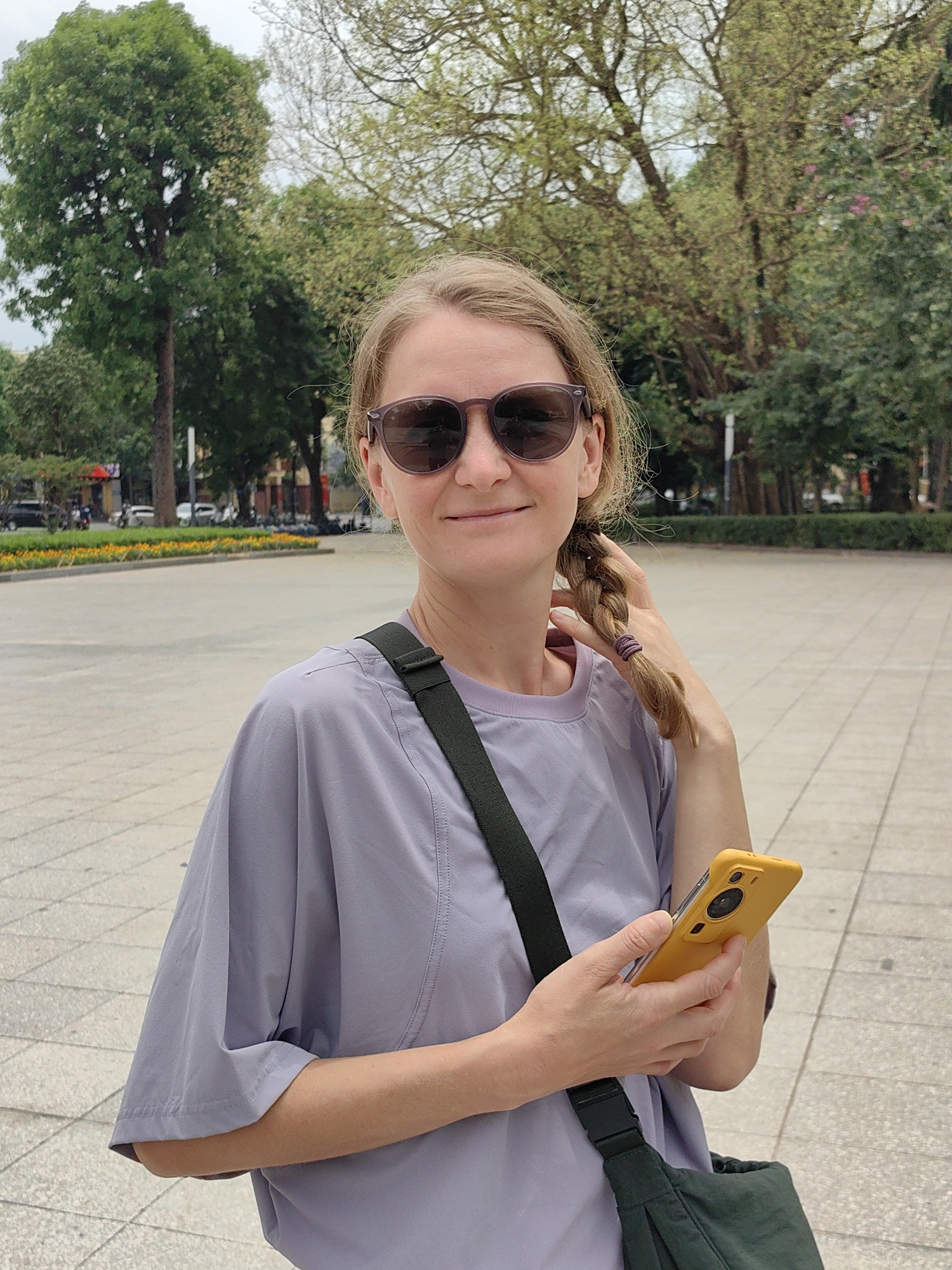 An outdoor, mid-shot portrait features a woman standing in a paved public space, likely a park or plaza in Hà Nội, Vietnam, during daytime with overcast skies. She looks towards the camera with a subtle smile, wearing dark sunglasses and a light purple, loose-fitting t-shirt. Her blonde hair is braided and draped over her left shoulder, secured with a purple elastic. A dark green cross-body bag is slung across her body. In her left hand, she holds a bright yellow smartphone, which has a prominent circular camera module on its back. She is casually touching her braid with her right hand. The paved ground beneath her consists of light-colored square tiles. The background reveals abundant green trees and foliage, beyond which lies a street bordered by a strip of yellow and orange flowers. Further down the street, blurred vehicles, likely motorbikes or scooters, and urban buildings are visible. The overall mood is relaxed and casual. There is no visible text in the image.