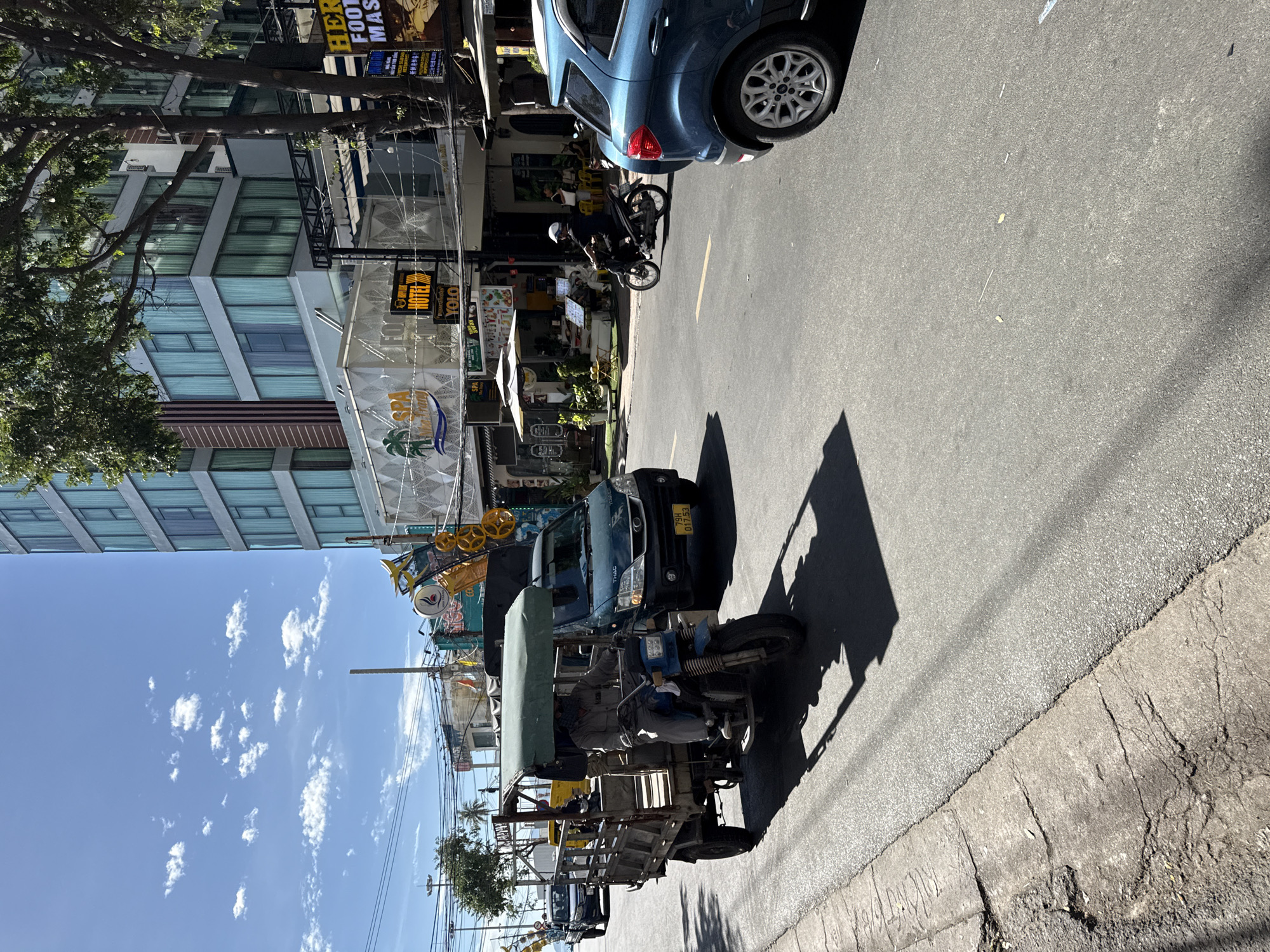 The image depicts a street scene in Nha Trang, Vietnam, under a clear blue sky with scattered clouds. A blue car is parked on the right side of the road. In the foreground, a three-wheeled vehicle, driven by a person wearing a mask and a grey uniform, is in motion. Behind it, a dark green van is also on the road. Buildings line the street, including a modern, multi-story building with many windows and several businesses with awnings and signage, some of which are in Vietnamese and English. One sign reads "SPA" with a palm tree graphic, another says "HOTEL YOLO." A person on a motorcycle is visible further down the street. Power lines are strung across the sky. The street itself is paved, with a cracked concrete sidewalk on the left. The overall impression is of a busy urban environment.