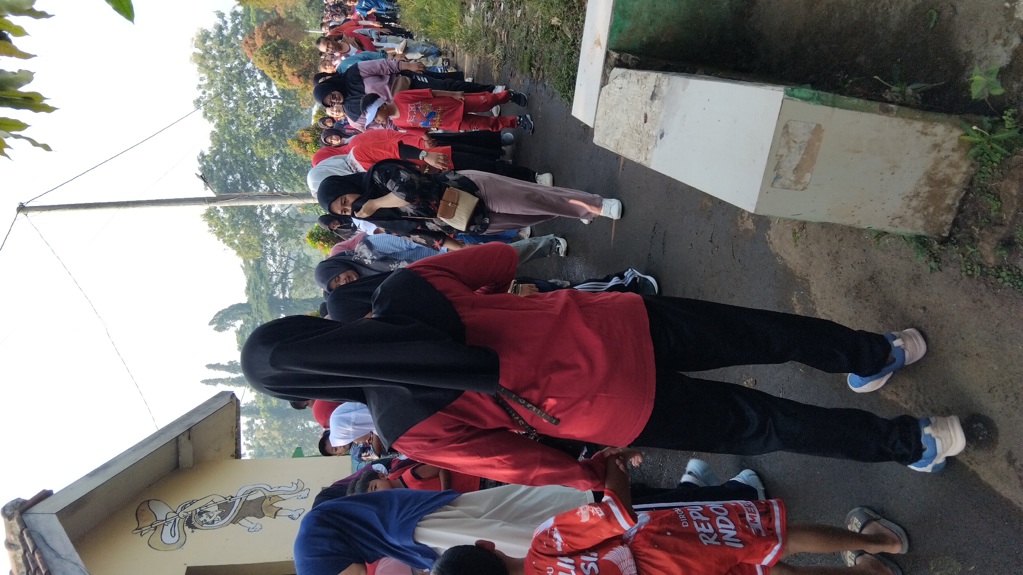 The image shows a group of people, likely participating in an outdoor activity or event, walking along a paved path next to a drainage ditch. Many individuals are wearing Islamic head coverings (hijabs), indicating a cultural context. The group appears to be moving from left to right within the frame. In the foreground, a child in a red shirt and shorts is visible. Behind them, an adult in a red shirt and black pants is walking. Further back, more people, including children and adults, are seen walking in a line. The environment includes a building with a mural on the left, trees and vegetation in the background, and a utility pole with wires. The lighting suggests it is daytime, possibly morning or late afternoon, with the sun casting a warm glow. The scene is consistent with a community gathering or procession in a semi-urban or suburban setting. The presence of Indonesian text on the child's shorts ("REPUBLIK INDONESIA") confirms the location as Indonesia. The overall atmosphere appears to be one of shared activity and movement.