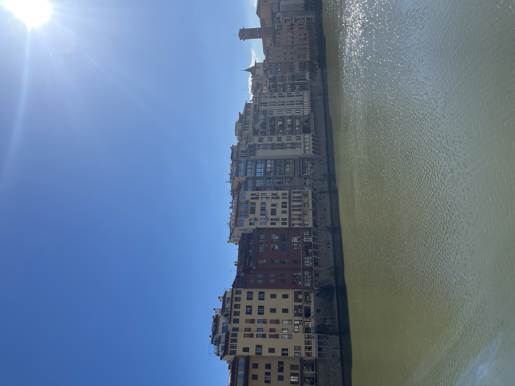 The image shows a daytime view along the Arno River in Florence, Italy. A row of multi-story buildings lines the riverbank. The buildings vary in color, including shades of yellow, beige, red, and gray. The sky is clear and blue with a bright sun at the upper left. The Arno River is a murky green color.