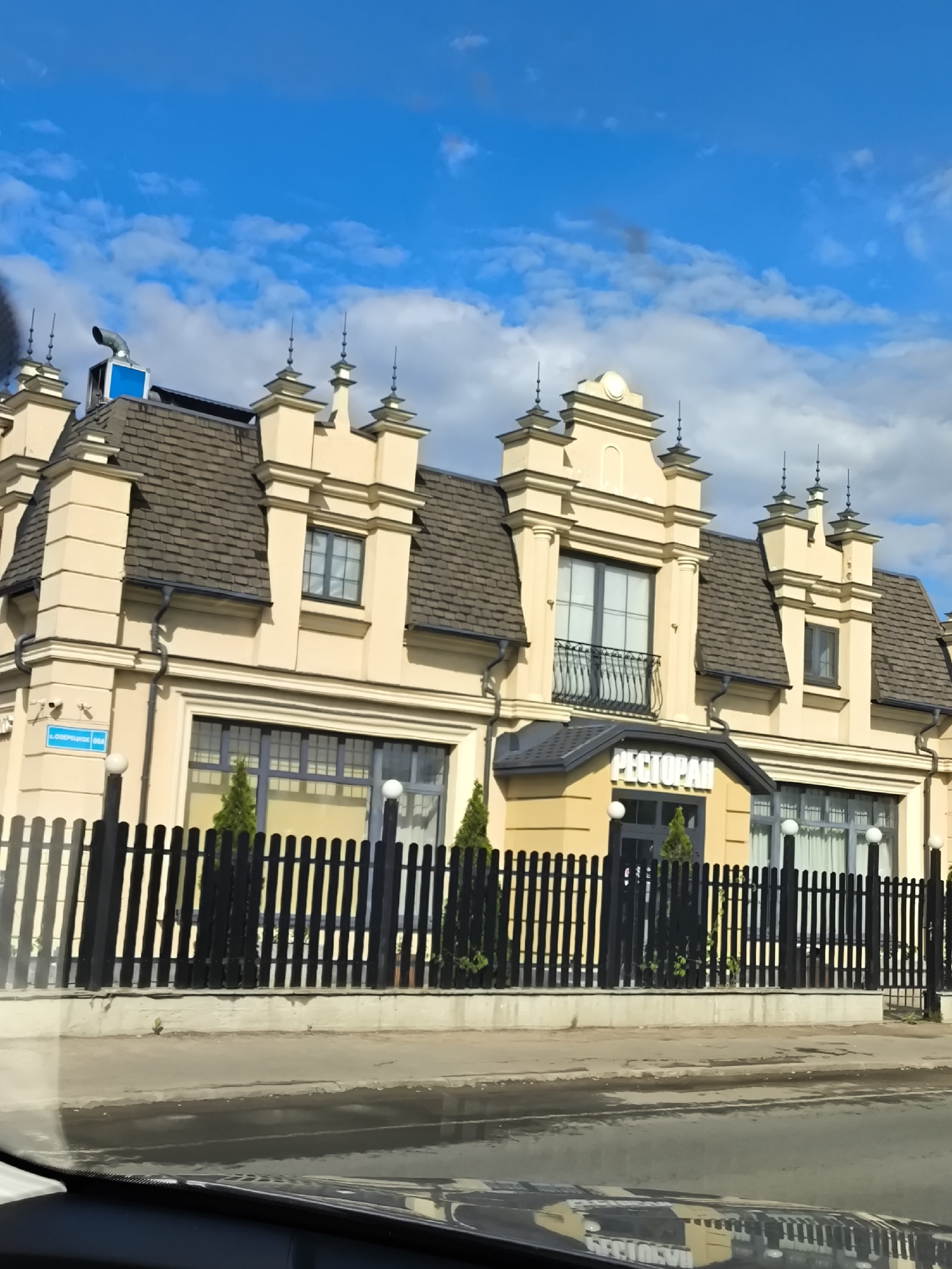 The image shows a restaurant in an urban setting. The restaurant building is cream-colored with a dark brown roof and is adorned with decorative architectural details. A black picket fence surrounds the front of the property, with small evergreen trees planted behind the fence. The street in front of the restaurant appears wet, and the scene is viewed from inside a vehicle. The sky above is blue with scattered clouds. A sign that reads "PECTOPAH" is visible above the entrance to the restaurant. A small blue sign that reads "L. O3ЕРЯНСЬКА 86А" is visible to the left of the building.