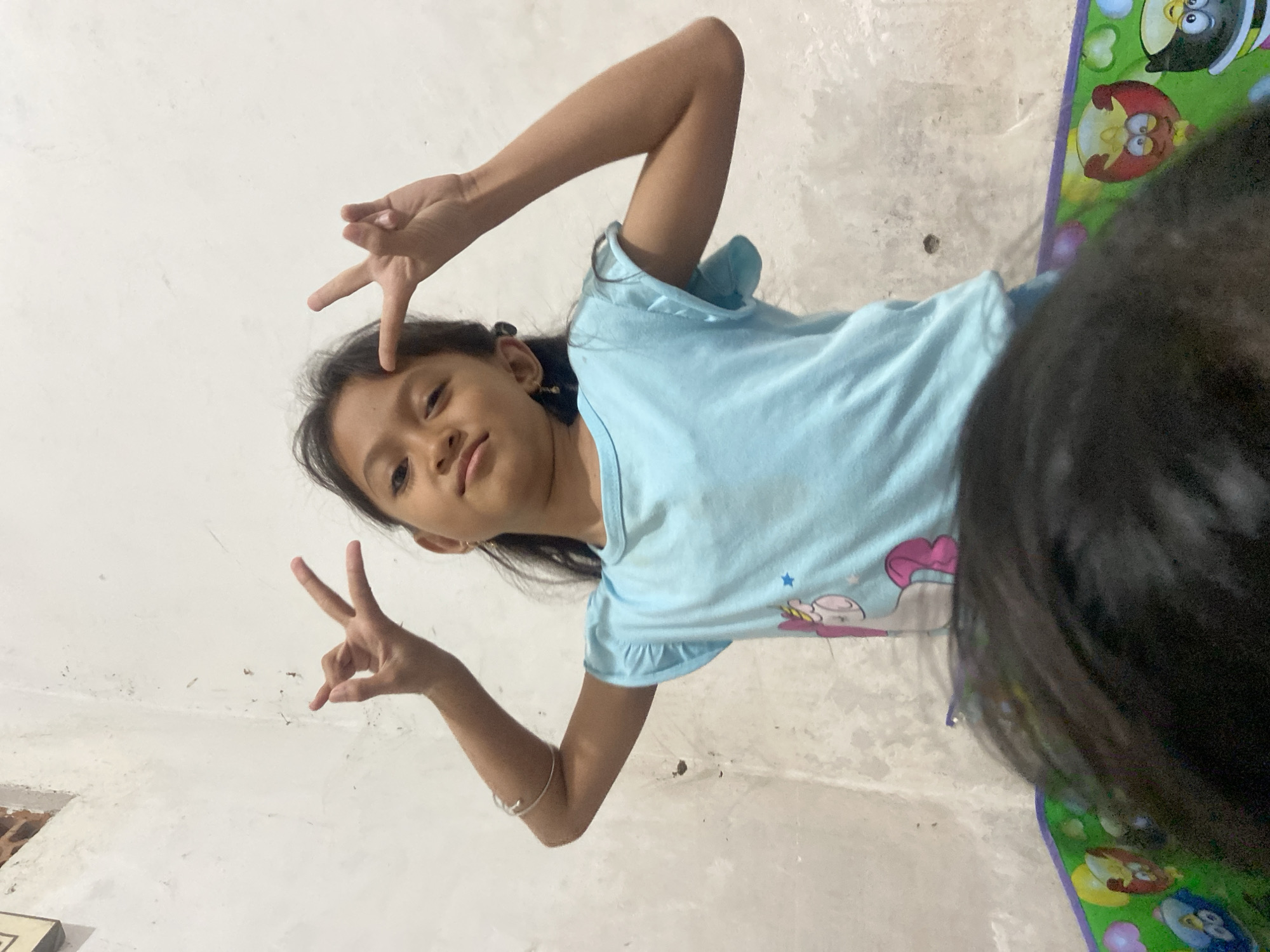 A young girl, likely of Indonesian descent given the location context of Baron, Indonesia, is pictured in a brightly lit indoor setting. She is wearing a light blue t-shirt adorned with cartoon characters. The girl has dark hair and is making a "peace sign" with both hands, one held near her forehead and the other extended outwards. Her expression is playful and engaging.
The background consists of a textured wall and a colorful mat featuring cartoon characters, possibly from a children's play mat. The lighting suggests daytime. The overall scene conveys a sense of casual indoor activity, with the girl posing for the camera. There is no visible text in the image.