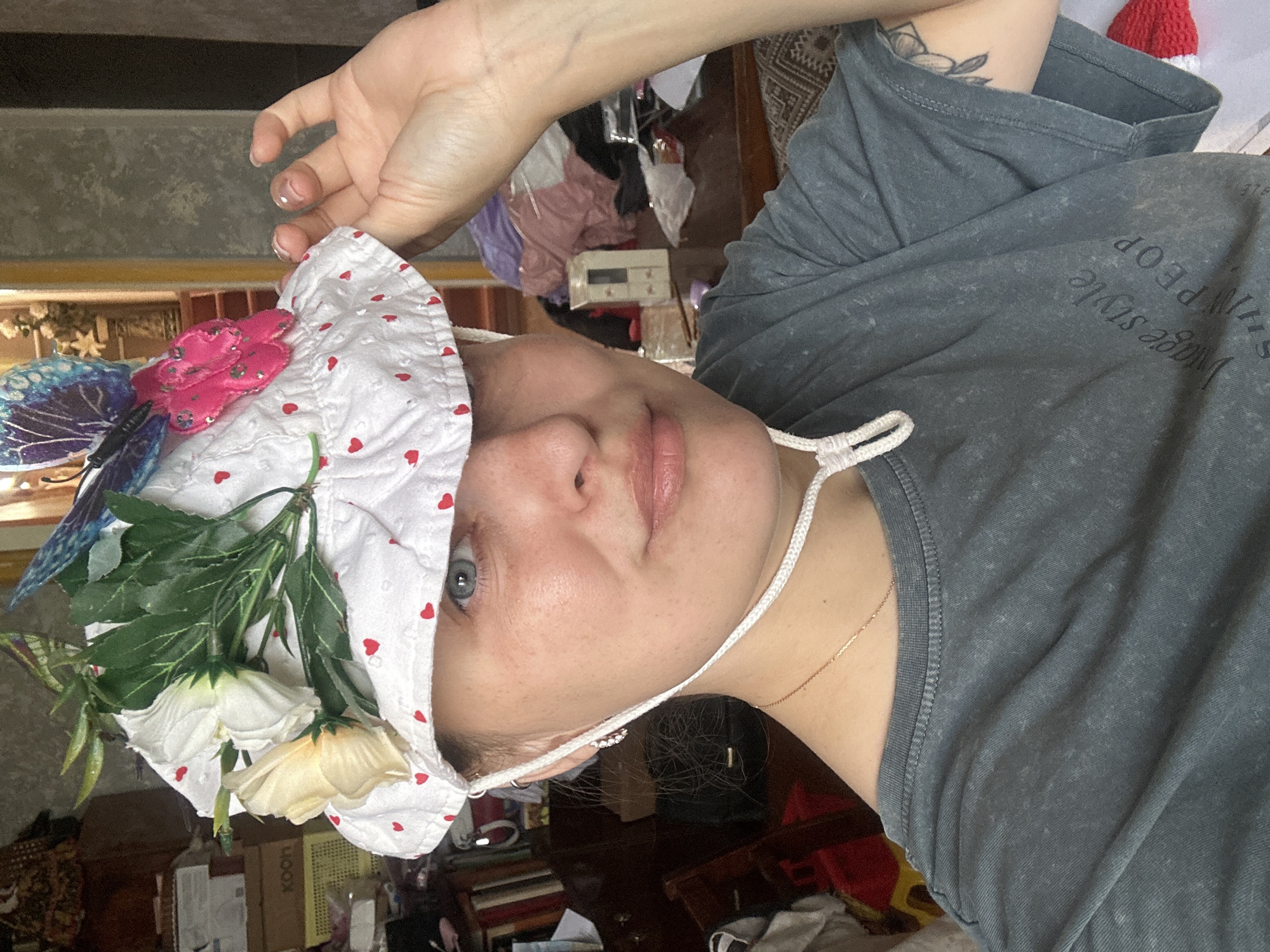 The image shows a woman wearing a white bucket hat decorated with artificial flowers and butterflies. The hat has a brim with a pattern of small red hearts. The woman has blue eyes and is looking at the camera. She is wearing a grey t-shirt with text printed on it. Her right hand is raised, holding the brim of the hat. The background appears to be an indoor setting, possibly a room in a house, with shelves filled with books and other items visible. There is also a glimpse of furniture and a window. The lighting suggests it is daytime. The location of Yaroslavka, Russia, is not explicitly indicated by any visual cues in the image.