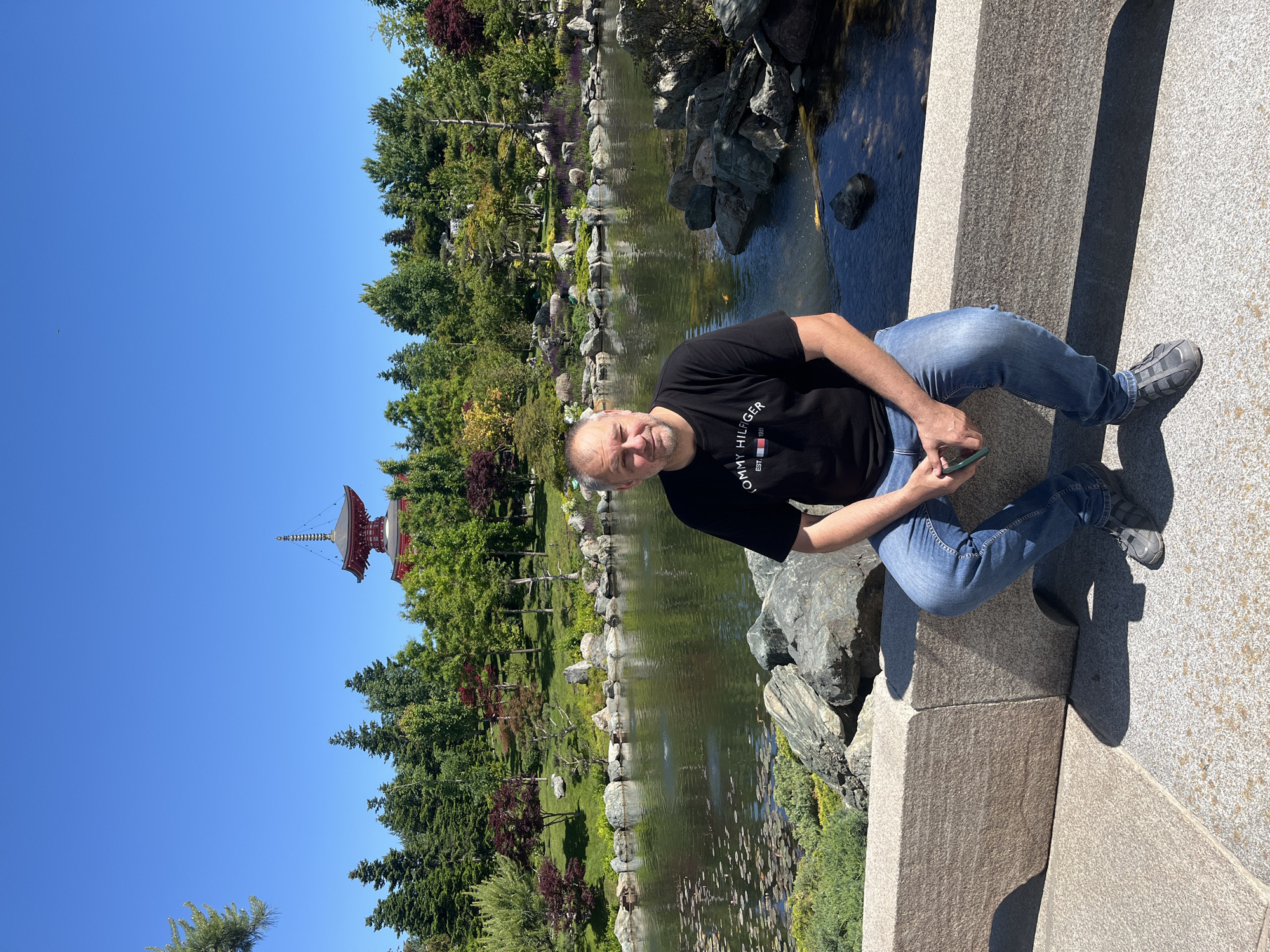 The image, correctly oriented, shows a man sitting on a stone ledge in a Japanese garden setting on a bright, sunny day. The man, with short, graying hair and a slight smile, is wearing a black T-shirt with "TOMMY HILFIGER" printed on it, blue jeans, and gray and black sneakers. He is holding a green-cased smartphone in his left hand.
He is positioned on the right side of the frame, looking directly at the viewer. To his left and behind him is a serene pond or small lake, reflecting the clear blue sky and bordered by carefully arranged rocks and lush green and reddish-purple foliage. Small lily pads dot the water's surface, and hints of fish are visible in the clear water.
In the far left background, a distinctive multi-tiered red pagoda-like structure with an ornate golden spire stands tall against the vibrant, cloudless blue sky. The surrounding landscape is rich with various trees and shrubs, creating a well-maintained park environment. Bright sunlight casts clear shadows on the stone ledge and the man, indicating midday or early afternoon. The scene evokes a tranquil atmosphere, consistent with the Japanese Garden in Galitsky Park, Krasnodar, Russia.