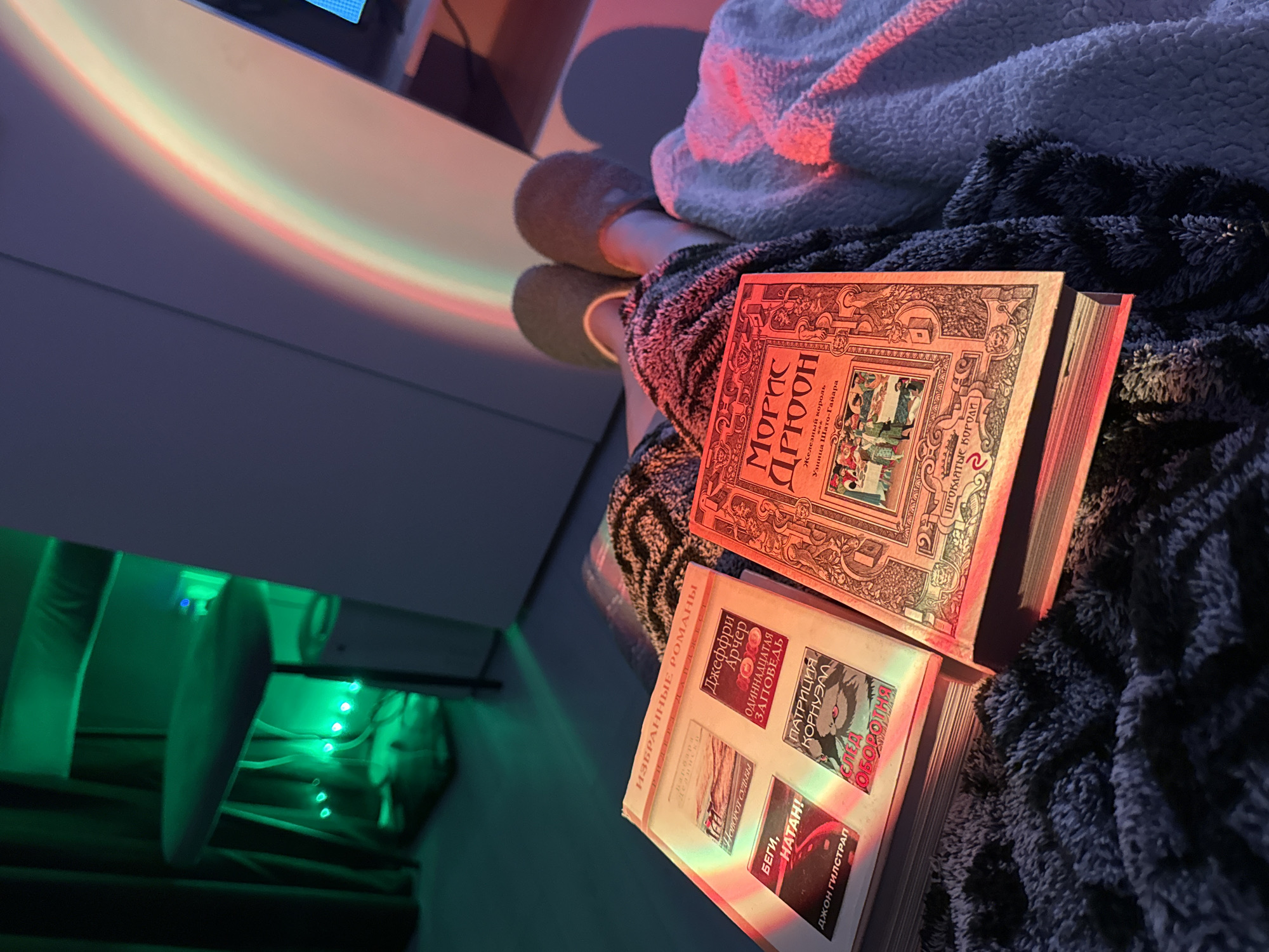 The image shows a person wearing slippers and covered with a blanket, reading two Russian books. The books are "Maurice Druon. The Iron King" and "Selected Novels. The Eleventh Commandment. Patricia Cornwell." A red-orange light illuminates the book on top. There is a shelf on the left side of the picture, with green LED lights underneath.