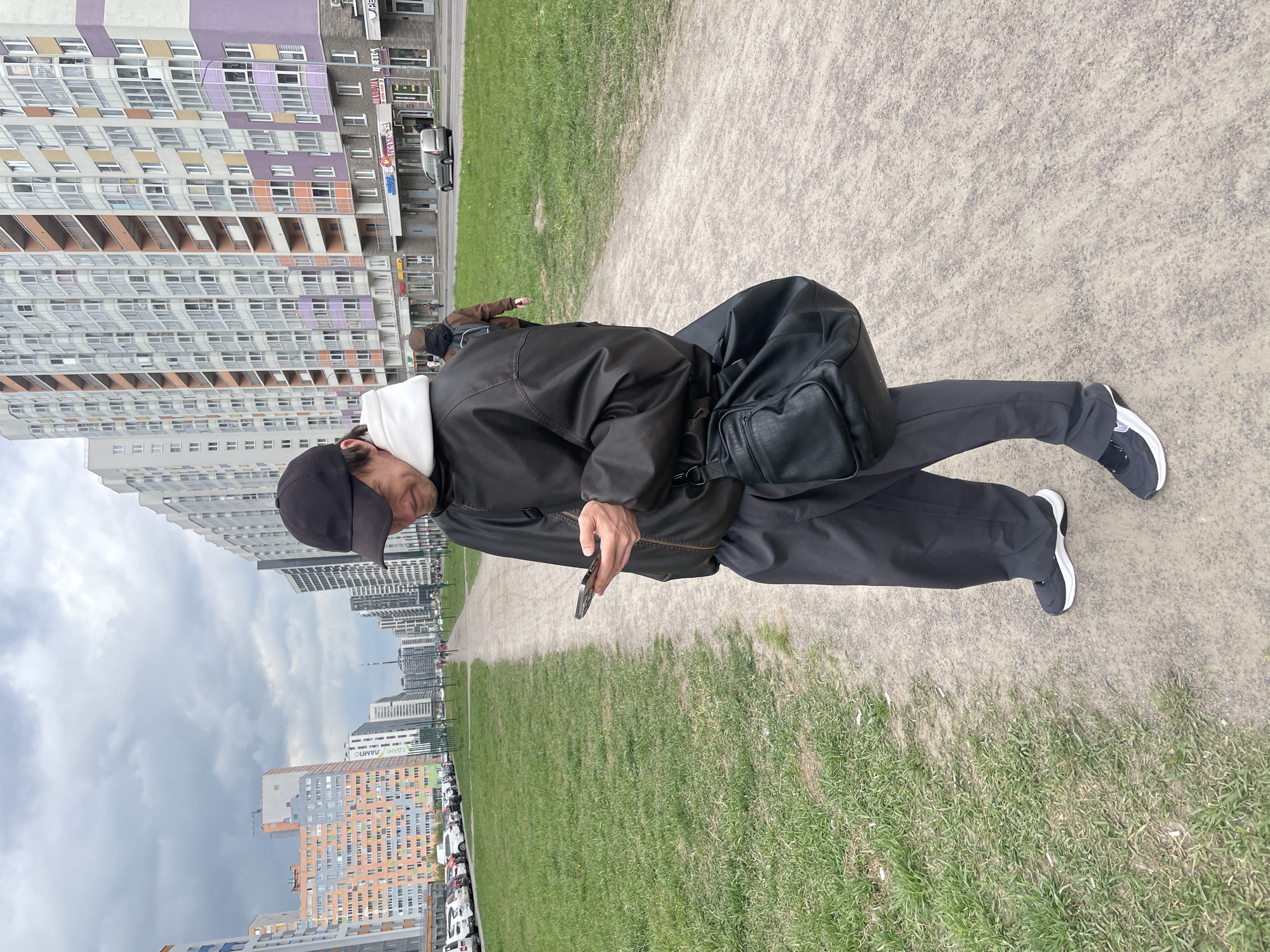 A person stands on a dirt and grass path in a city park. The person wears a black jacket, black pants, black shoes with white trim, and a black baseball cap. The person is looking down at a smartphone in their hand. A black bag hangs on their shoulder. In the background are tall apartment buildings and parked cars. The location is a park in the city of Unknown, Russia.