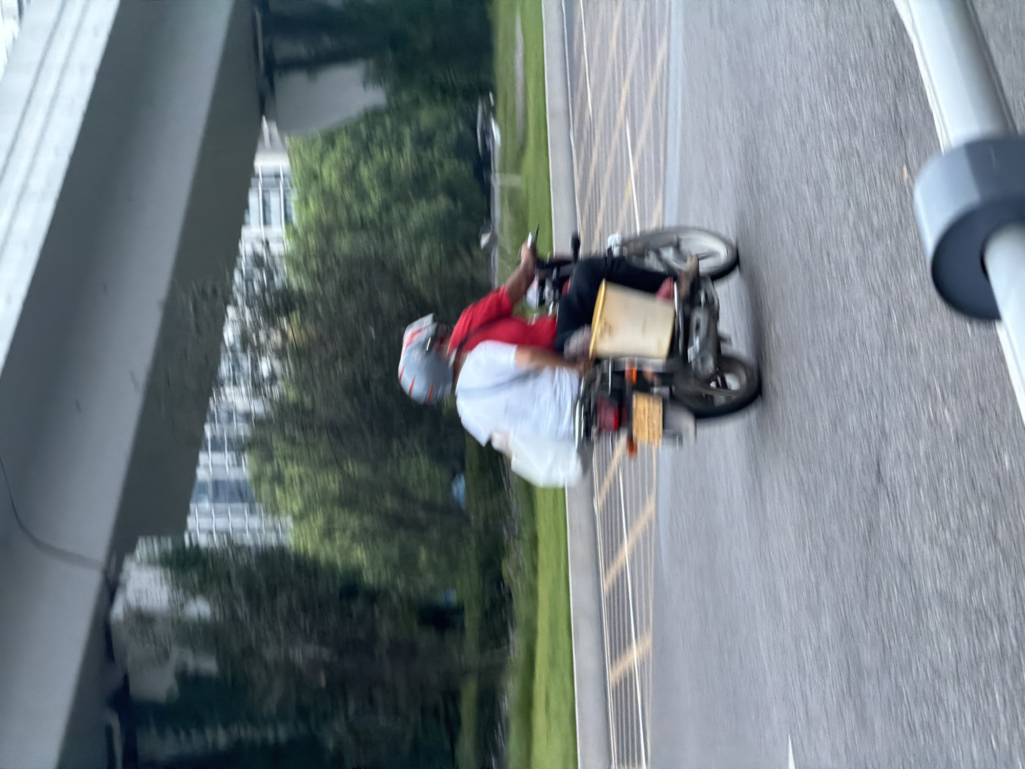 A medium shot captures two individuals riding a motorcycle on a city street in Jiangjin, China. The person in the front is wearing a red shirt and a helmet, while the person in the back wears a white shirt and a helmet. Both individuals are blurred due to motion. A white bag is slung over the shoulder of the person in the back. A yellow bucket is attached to the side of the motorcycle, and a license plate is visible at the rear. The motorcycle is in motion on an asphalt road with lane markings. In the background, trees and buildings are visible, with an elevated concrete structure overhead. The overall scene is slightly blurred, suggesting the photograph was taken while in motion.