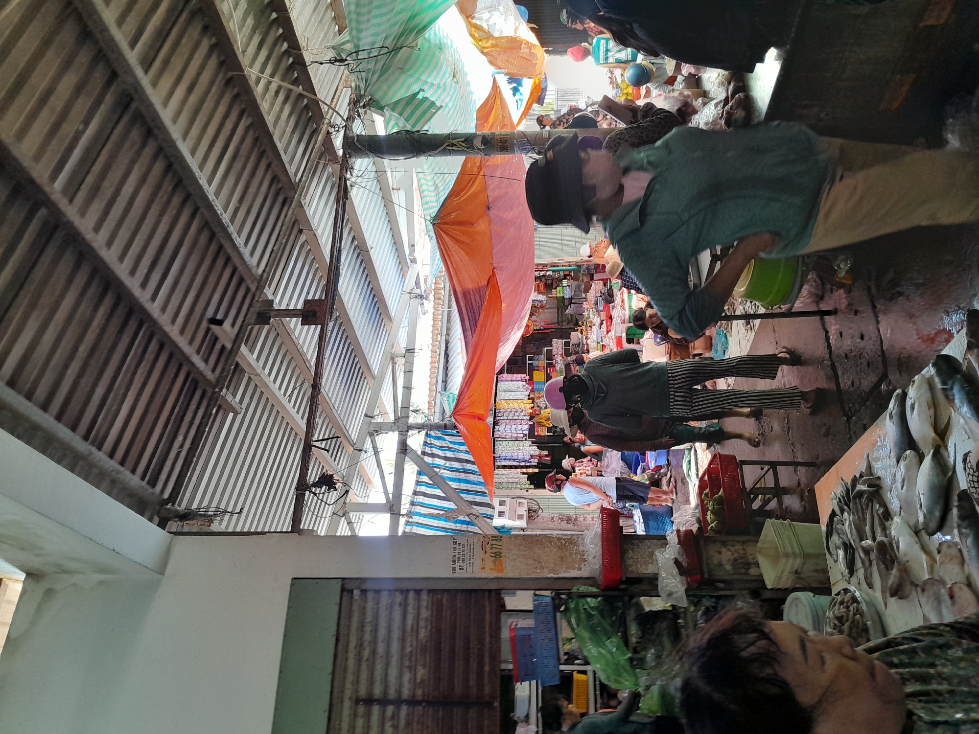 The image depicts a bustling outdoor market, likely a fish market, in Phan Thiet Ward, Vietnam. The market is covered by a corrugated metal roof supported by beams, with additional tarps and awnings in various colors, including orange, green, and blue and white stripes, providing shade. The ground appears wet, suggesting it may have recently rained or is part of the market's operational environment.
Several individuals are present, engaged in buying, selling, or transacting business. In the foreground, a woman with dark hair is partially visible, looking down at a table laden with fresh fish. Another woman in a green hooded jacket and light-colored pants stands with her hands on her hips, facing away from the camera. Behind her, other people are milling about, some wearing hats.
The market stalls are filled with products, prominently featuring an abundance of fresh fish displayed on tables. Crates and baskets are visible, some containing produce or other goods. A sign with Vietnamese text is affixed to a pillar on the left side of the image, possibly advertising a business or providing market information. The overall atmosphere is one of a busy marketplace with goods laid out for sale.