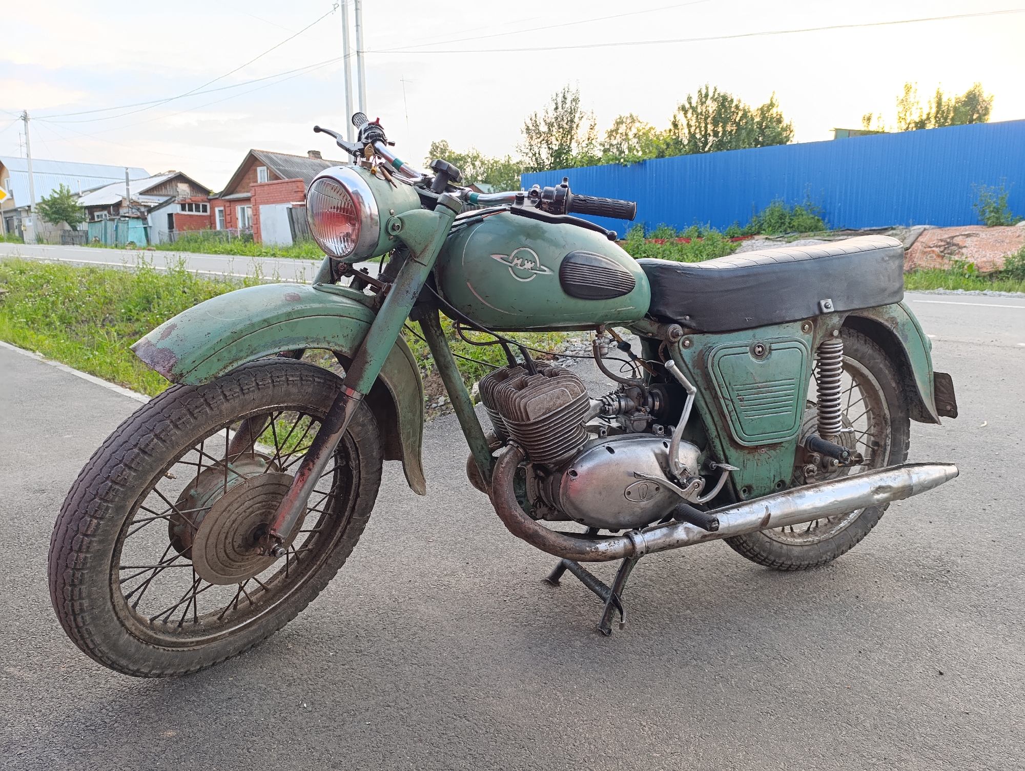 A vintage green motorcycle, identified as a motorcycle produced in Irbit, Russia, is parked on an asphalt road. The motorcycle is positioned at an angle, with its front wheel closer to the viewer. It is a two-wheeled vehicle with a visible engine, exhaust pipe, seat, handlebars, and a prominent round headlight. The motorcycle appears weathered, with signs of rust and wear.
In the background, a rural or semi-rural landscape is visible. There are several houses with varying architectural styles and colors, some appearing older and in need of repair. A blue corrugated metal fence runs along the right side of the background, separating the road from what appears to be overgrown vegetation and some construction materials. The sky is overcast with hints of sunset light filtering through. The scene suggests an outdoor setting, possibly a residential area in or near the city of Irbit, Russia, given the implied origin of the motorcycle. No people are present in the image.