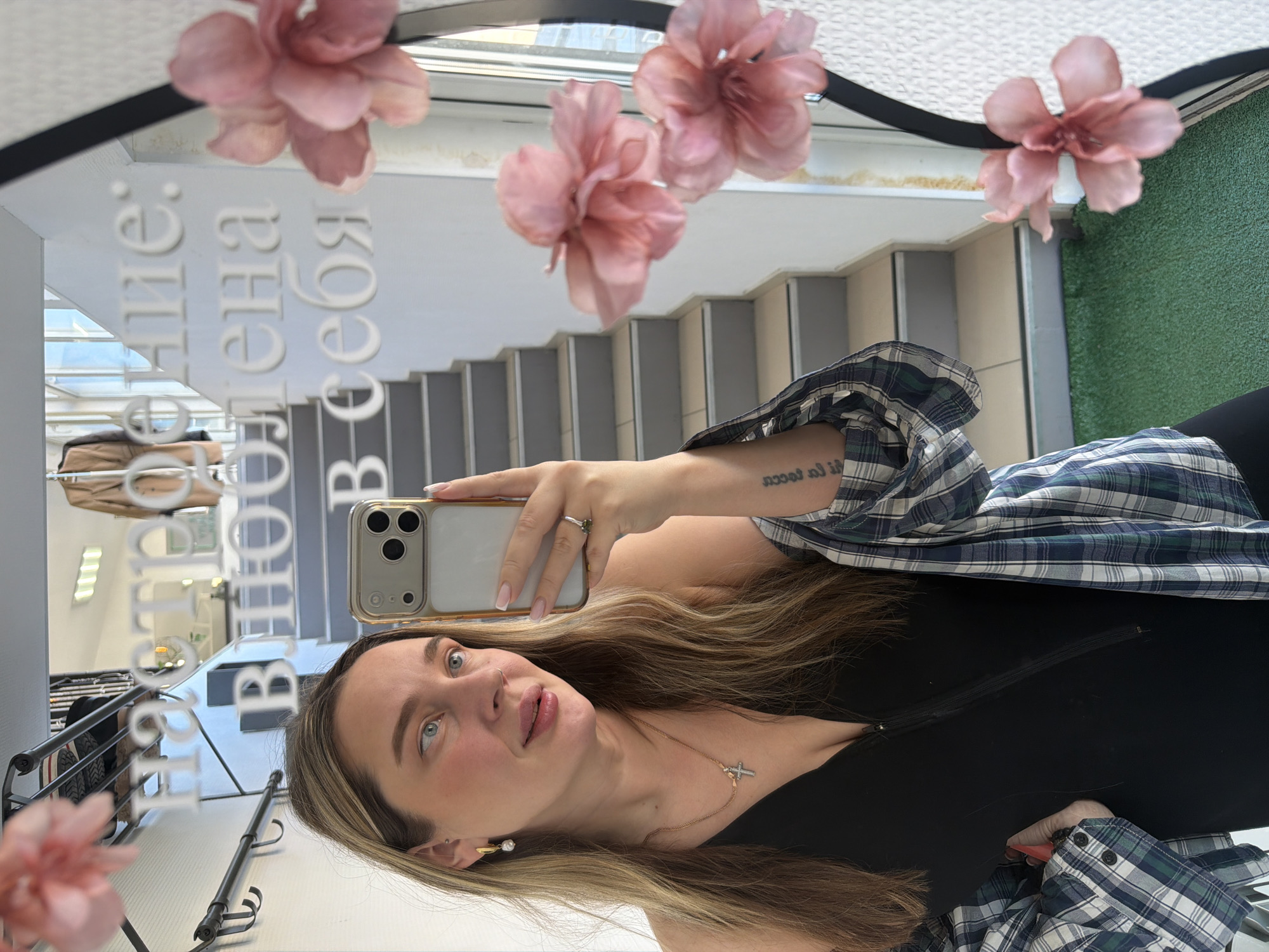 A young woman is taking a selfie in a mirror. The mirror is decorated with pink flowers and has text on it, likely in Russian. The reflection shows a staircase, a clothing rack with garments, and potentially a patch of artificial turf. The woman is wearing a black top and a plaid shirt. She has a tattoo on her arm and is holding a gold-colored iPhone. Based on the information from the previous turn, the location is presumed to be in the city of Chaikovsky, Russia.