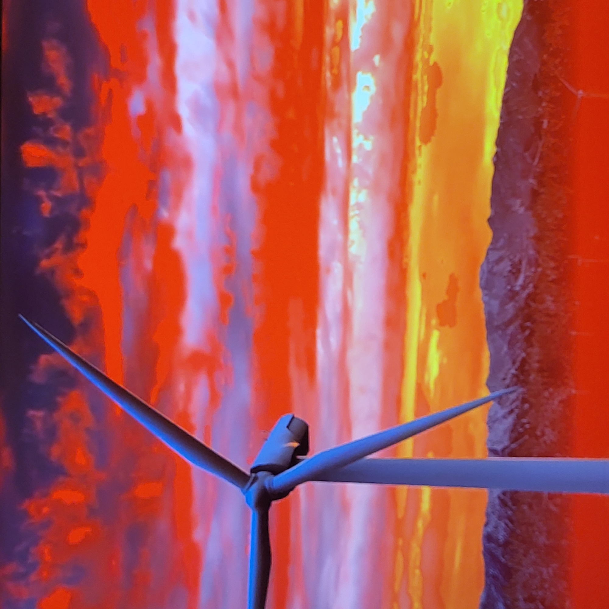 The image depicts a wind turbine set against a vibrant, abstract background of swirling colors. The background features a blend of red, orange, yellow, and purple hues, suggestive of a sunset or fire-like effect. The turbine itself is rendered in shades of blue and gray, standing out against the warm-toned backdrop. Given the location of Yerevan, Armenia, it's possible the turbine is part of a renewable energy initiative within the city or its surrounding areas. The image's artistic style and dramatic color palette suggest a promotional or illustrative purpose, rather than a purely documentary one.