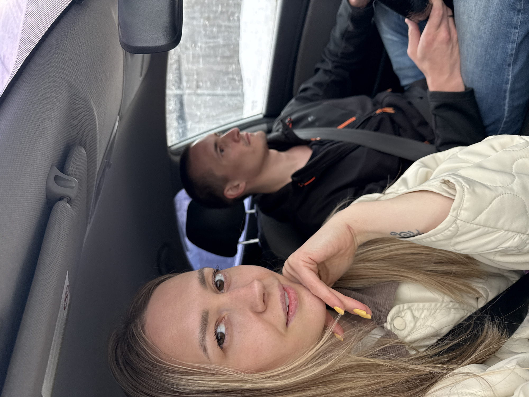 Two people are in a car. A woman with blonde hair and a white jacket is taking a selfie, with her hand on her face. A man is behind her with a black jacket and jeans. They are both wearing seatbelts. It appears to be raining outside, as droplets are visible on the car window. The location is potentially the city of Chaikovsky, Russia.