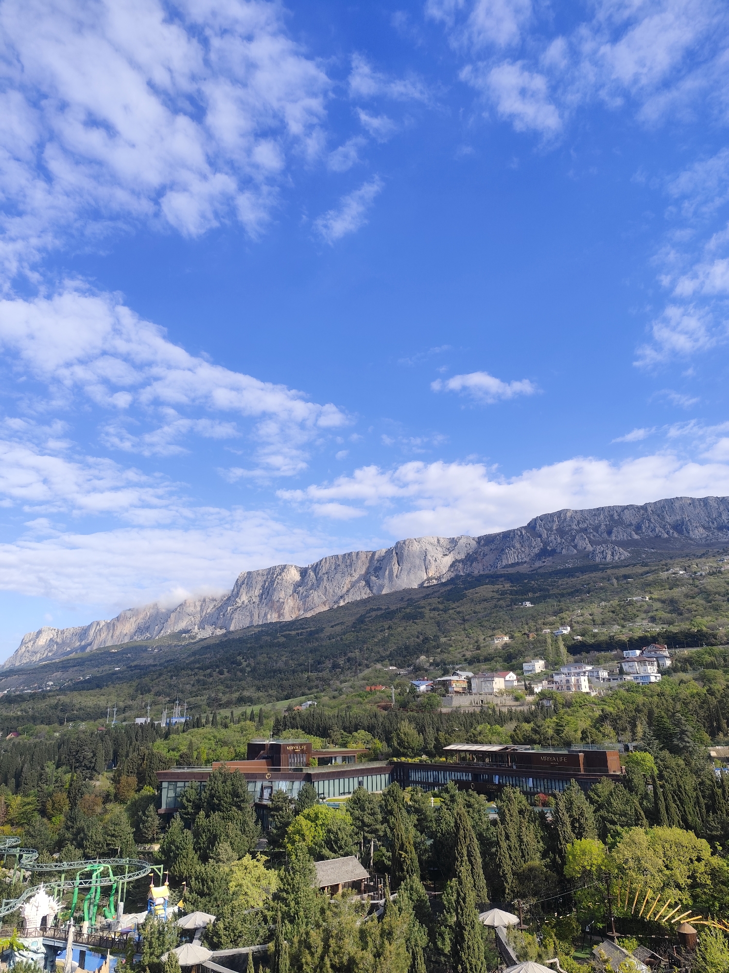 The image depicts a landscape in Симеизский поселковый совет, Ukraine, with a prominent mountain range in the background. The mountains are mostly bare rock with patches of green vegetation. In the foreground, there are trees, buildings, and other signs of civilization. A long, multi-story building with the name "Mriya Life" on its roof is visible. Below this building is a collection of trees and a visible amusement park. The sky is blue with scattered white clouds.