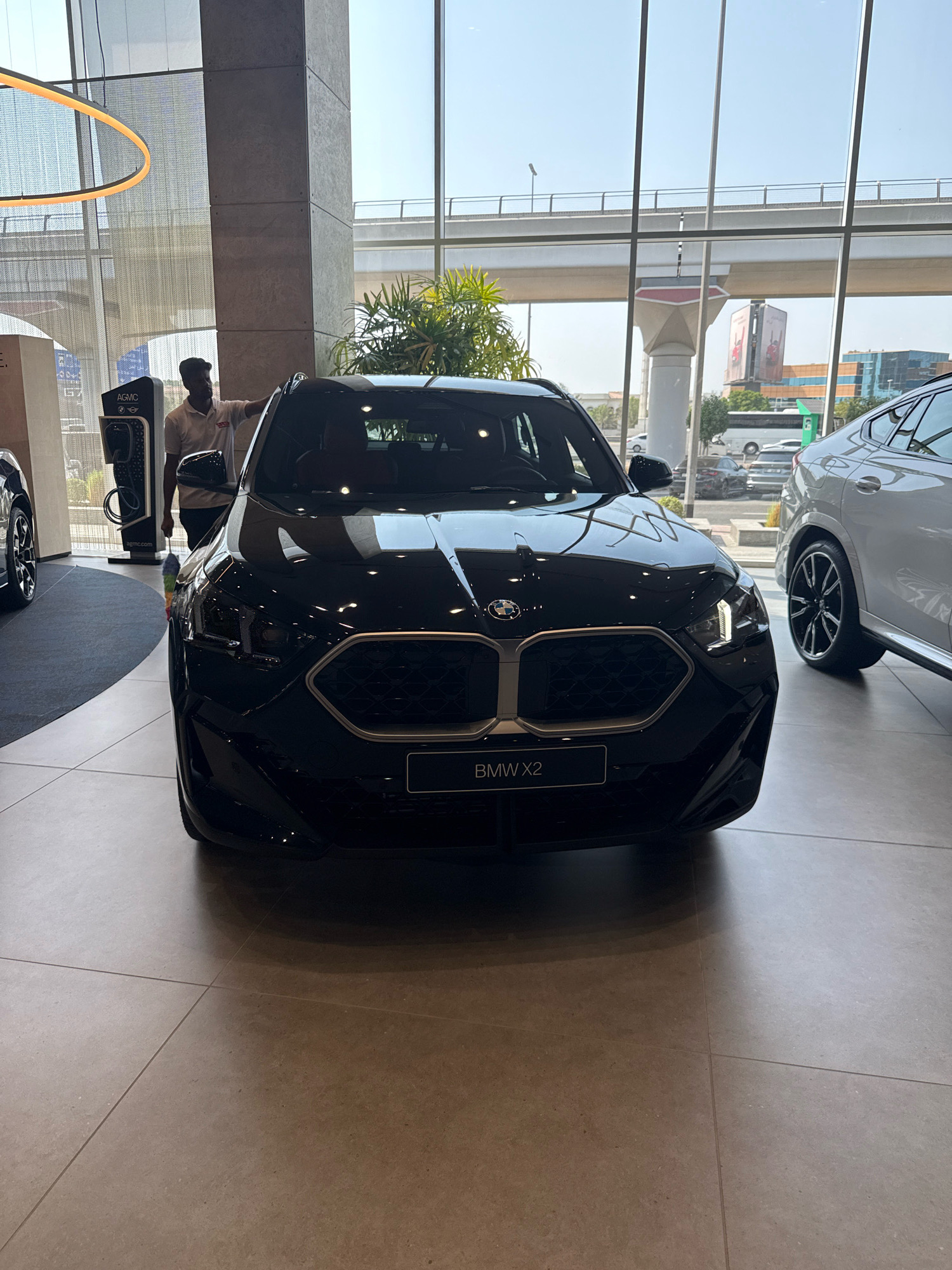 A black BMW X2 is parked in a showroom in Dubai, United Arab Emirates. A man in a white polo shirt and black trousers stands next to the car, holding a multi-colored duster. The showroom has a modern, open design with large windows and high ceilings. Outside the windows, an elevated highway structure and contemporary buildings are visible, suggesting an urban setting. The floor is tiled with large, light-colored tiles. Another car, a white BMW, is parked to the right of the black BMW.