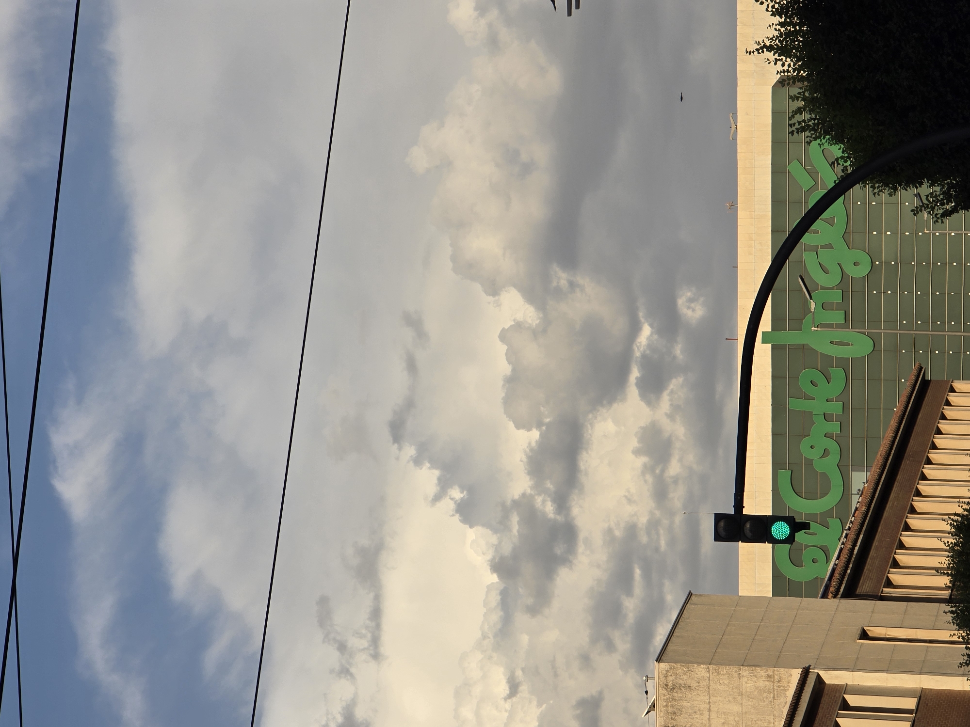 The image shows a street scene with a traffic light showing green, a building with a large green logo that reads "El Corte Inglés", and a partially visible building on the left. The sky is filled with dramatic clouds, with some areas of blue sky visible. Overhead, there are power lines crossing the frame. The overall lighting suggests it might be late afternoon or early evening. The scene appears to be in an urban environment.