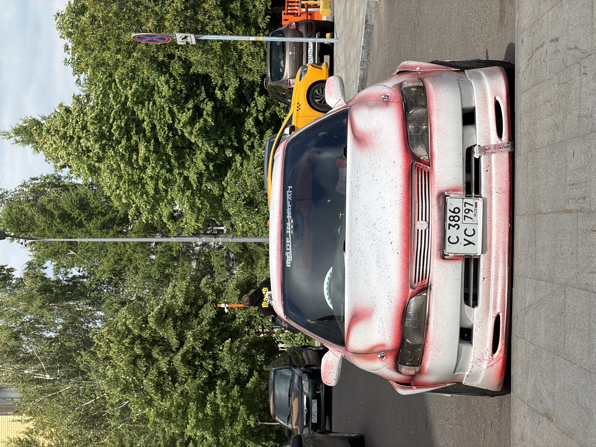 This image depicts a vibrant street scene in Moscow, Russia, during daylight hours. The primary subject is a highly customized white car with bold red spray-painted patterns, giving it a distinctive, possibly drift-inspired, aesthetic. Visible details on the car include a "MADE IN JAPAN" sticker with Japanese characters on the windshield, a "MIDNIGHT" label on a strap attached to the front bumper, and a Russian license plate displaying "C 386 YC 797 RUS". A person with dark hair, wearing a black hoodie with yellow text, is partially visible near the customized car's windshield.
Behind the customized vehicle, a bright yellow taxi cab is parked, followed by a dark grey car with a partially visible Russian license plate, "a 922 ex 97". Another dark car with the partially visible plate "x 585 cy 790" is parked further down the street. The setting features a row of abundant green trees, some with small yellow or white flowers, lining the street. A tall pole stands between the cars and trees, displaying two signs: a standard "No Stopping" sign (red circle with a blue cross) and an adjacent square sign with an icon resembling a surveillance camera. The cars are parked on a concrete sidewalk next to a paved road. The clear sky and bright illumination suggest good weather. The scene captures an urban environment with a mix of street life and greenery.