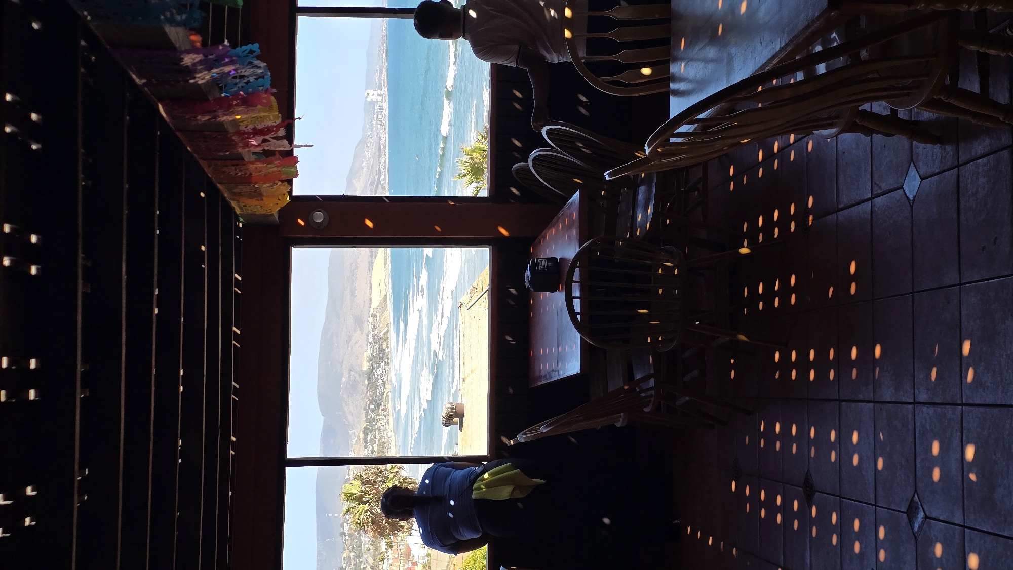 The image captures an interior view of a restaurant with a large window overlooking the ocean and coastline of Puerto Nuevo, Mexico. The foreground shows several wooden tables and chairs, with dappled light patterns cast on the tiled floor from an unseen light source, possibly decorative lights or sunlight filtered through a patterned structure.
Two individuals are visible. On the left, a person with dark hair pulled back is leaning against a dark surface, looking out the window. They are wearing a blue shirt and a yellow cloth is draped over their shoulder. On the right, a person facing away from the camera is standing near the window, also looking out at the sea. They are wearing a light-colored polo shirt.
The window reveals a scenic view of the Pacific Ocean with waves breaking on the shore. The coastline is dotted with buildings, and hills rise in the background under a bright sky. Colorful papel picado decorations are strung across the ceiling beams. The overall impression is that of a casual dining establishment with a prime ocean view.