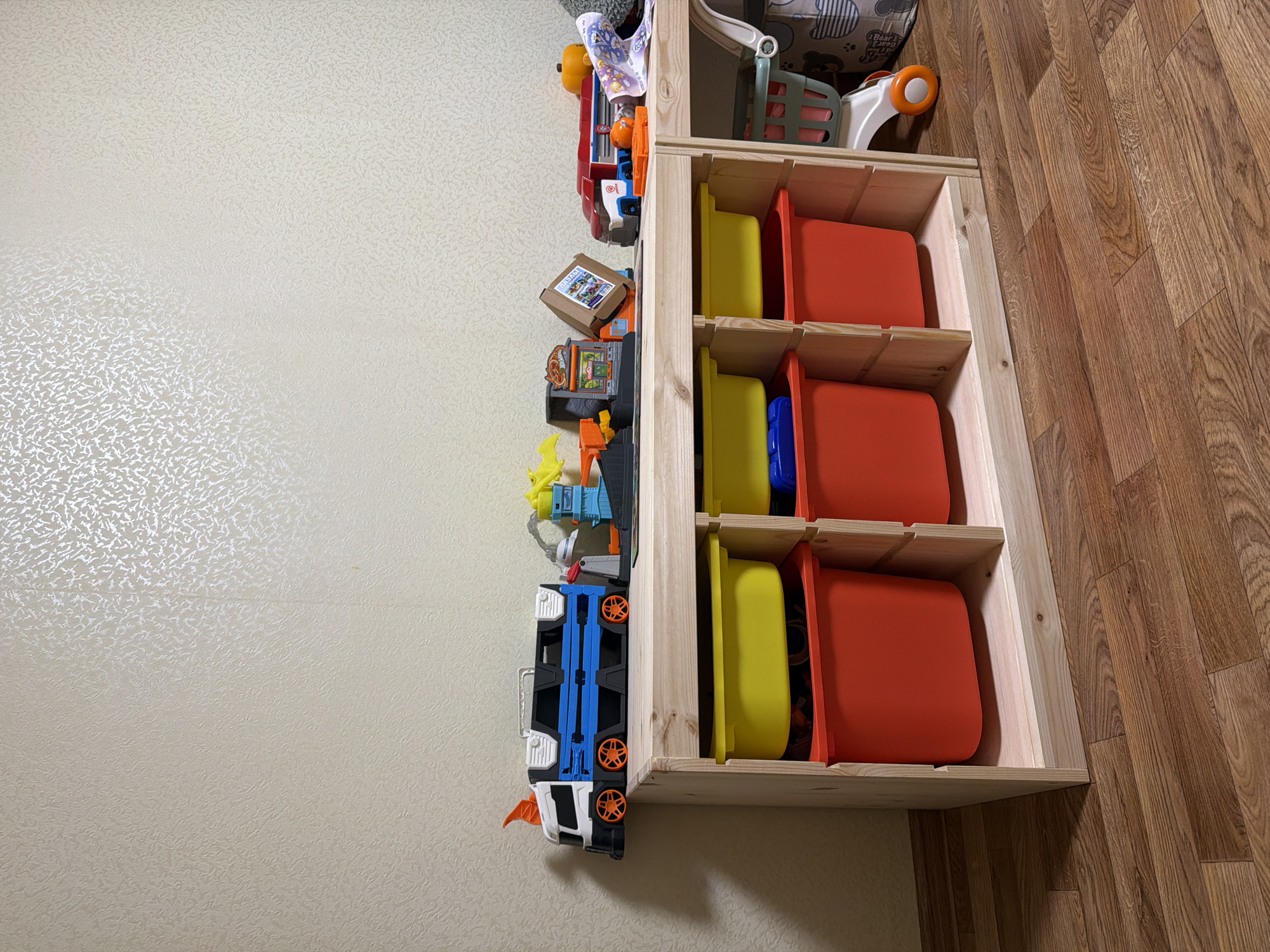 The image shows a wooden toy storage shelf against a textured beige wall. The shelf has three columns, each with two open cubbies. The top cubbies are filled with yellow plastic bins, and the bottom cubbies are filled with red plastic bins. On top of the shelf are various toys, including a large blue and white truck, a toy construction set with a yellow dinosaur, a red and white vehicle, a small orange pumpkin, and a folded patterned cloth. To the right of the shelf is a white toy shopping cart with green and orange accents. The floor is a wooden laminate with a brown, wood-grain pattern. The overall scene depicts a child's play area. The exact location in Yablonovskiy, Russia is not discernible from the image.