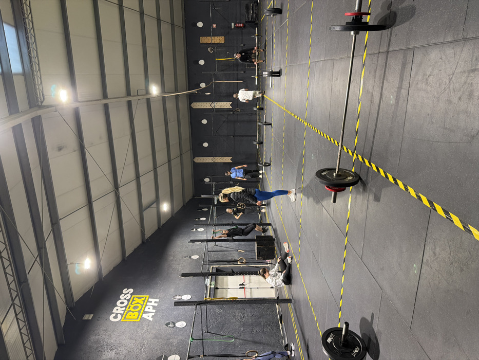 A brightly lit indoor gym shows several individuals spaced apart. They are engaged in various workout routines with equipment like pull-up bars and gymnastic rings. Barbells with weight plates are placed along the training area. Black and yellow hazard stripes run along the floor, serving as visual markers. "CROSS BOX APH" is written on the wall.