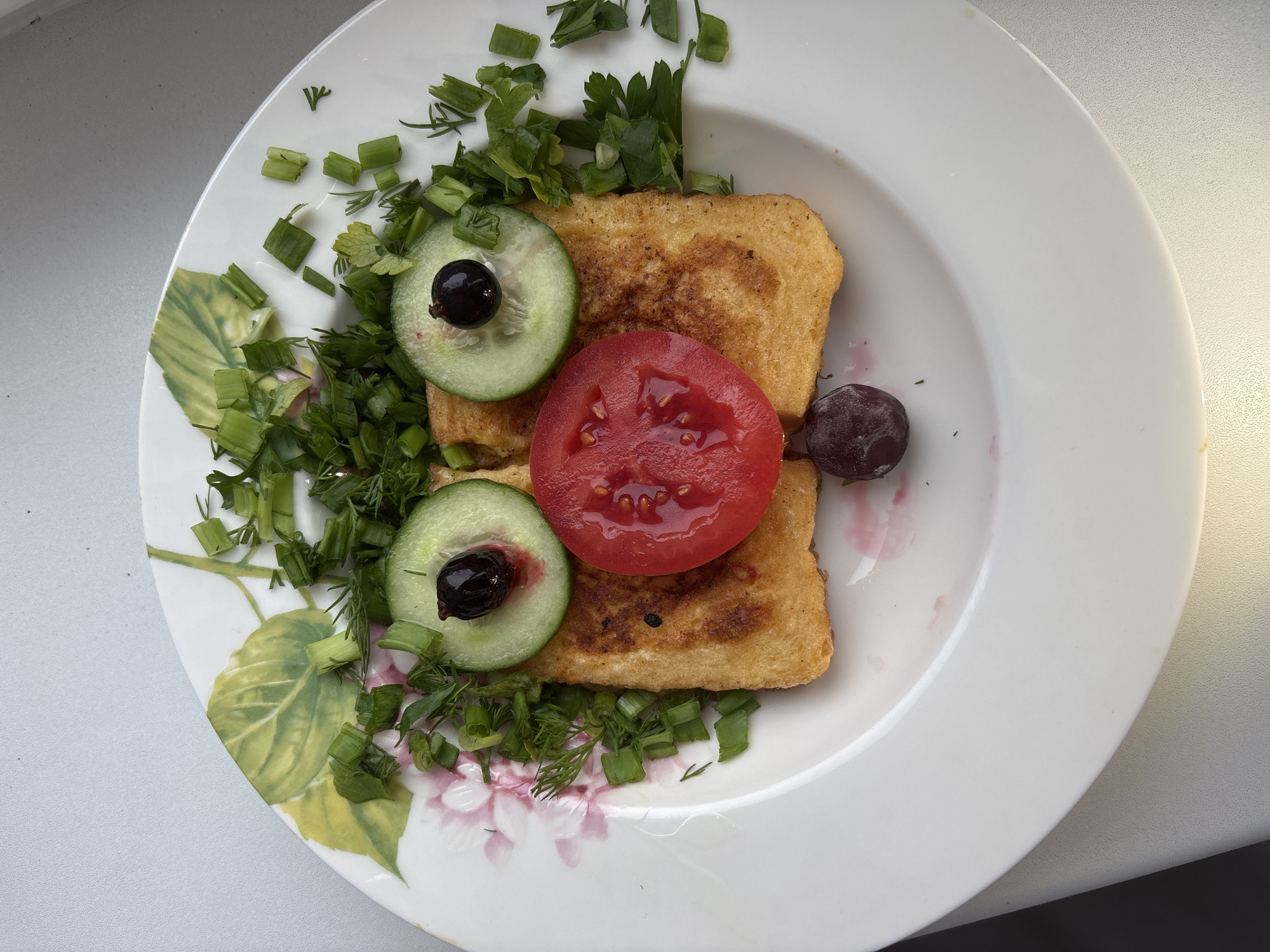 A top-down, full shot depicts a white, patterned plate containing a simple meal. The plate features floral designs in shades of green, pink, and white along its rim. The meal consists of two pieces of golden-brown toast, a slice of red tomato, two cucumber slices each topped with a dark berry, and loose chopped green herbs and green onions scattered around the plate. One additional dark berry is placed to the right of the tomato slice. The plate is set on a white surface. The location in Izhevsk, Russia is unknown.