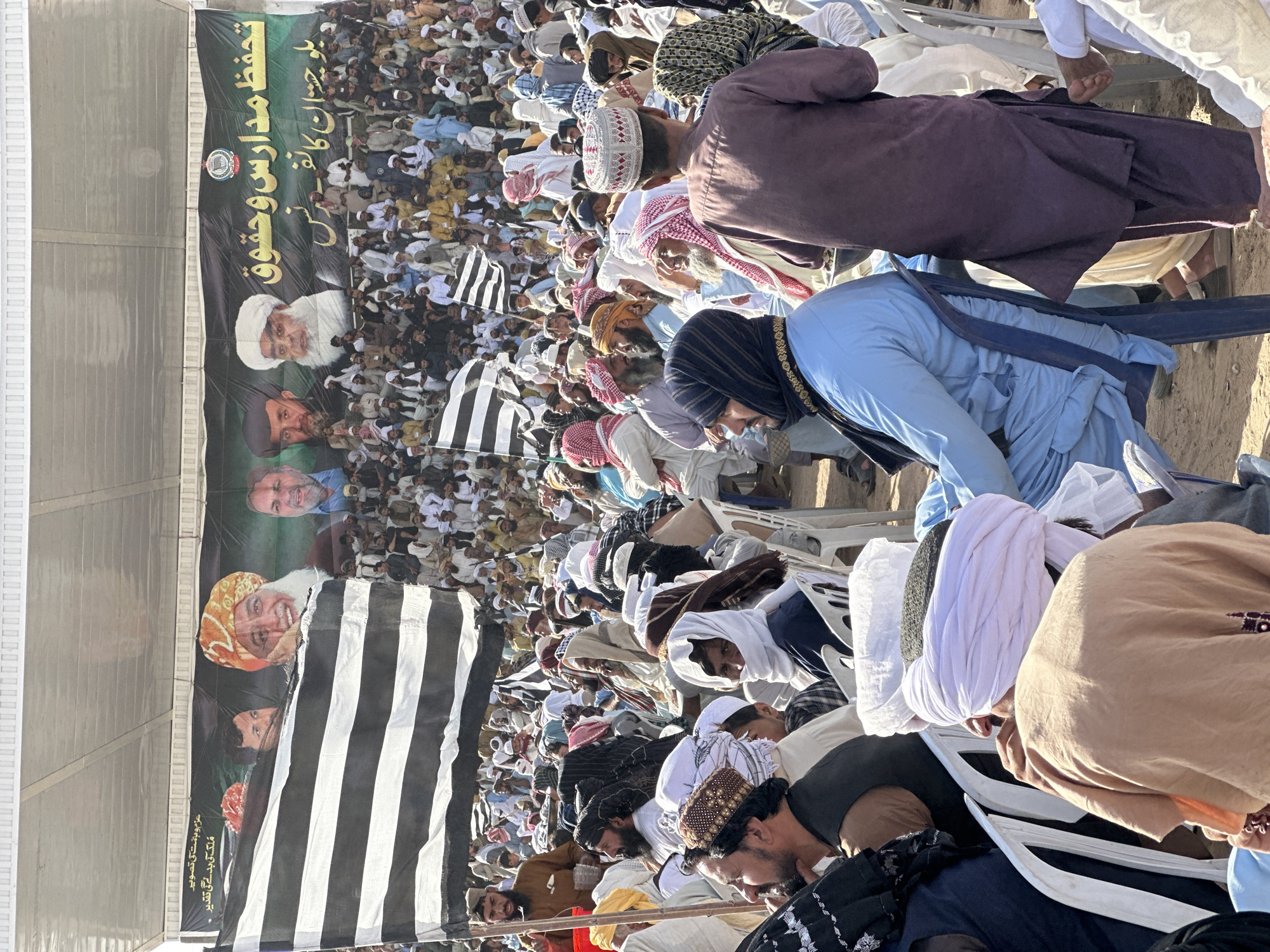 This outdoor image captures a large gathering of men, likely a political or religious conference, in Pishin, Pakistan. The scene is dominated by a diverse crowd, mostly adult men, dressed in traditional attire including kurtas, shalwar kameez, and various head coverings such as white, patterned, and solid-colored turbans or kufis. Many individuals are seated on white plastic chairs or kneeling/standing on the dusty ground, suggesting collective participation in an event.
A prominent dark green/black banner stretches across the upper left side of the frame. It features white text in Arabic/Urdu script and several portraits of men, including one with a white turban and beard, and another with a patterned orange head covering. The visible text on the banner reads: "Protection of Madrassas and Rights" (تحفظ مدارس وحقوق), "And Balochistan Conference" (و بلوچستان کانف- پرنس), along with slogans like "An image of determination and courage" (عزم و ہمت کی تصویر) and "The destiny of the country will change" (ملک کی بدلے گی تقدیر). A circular logo is also present.
Throughout the crowd, several black and white striped flags are visible, held by participants or standing upright. These flags are characteristic of the Jamiat Ulema-e-Islam (F) political party. The bright daylight indicates the event is taking place outdoors during the day under sunny, dry conditions. The overall impression is one of a large, organized gathering focused on religious and political discourse.
