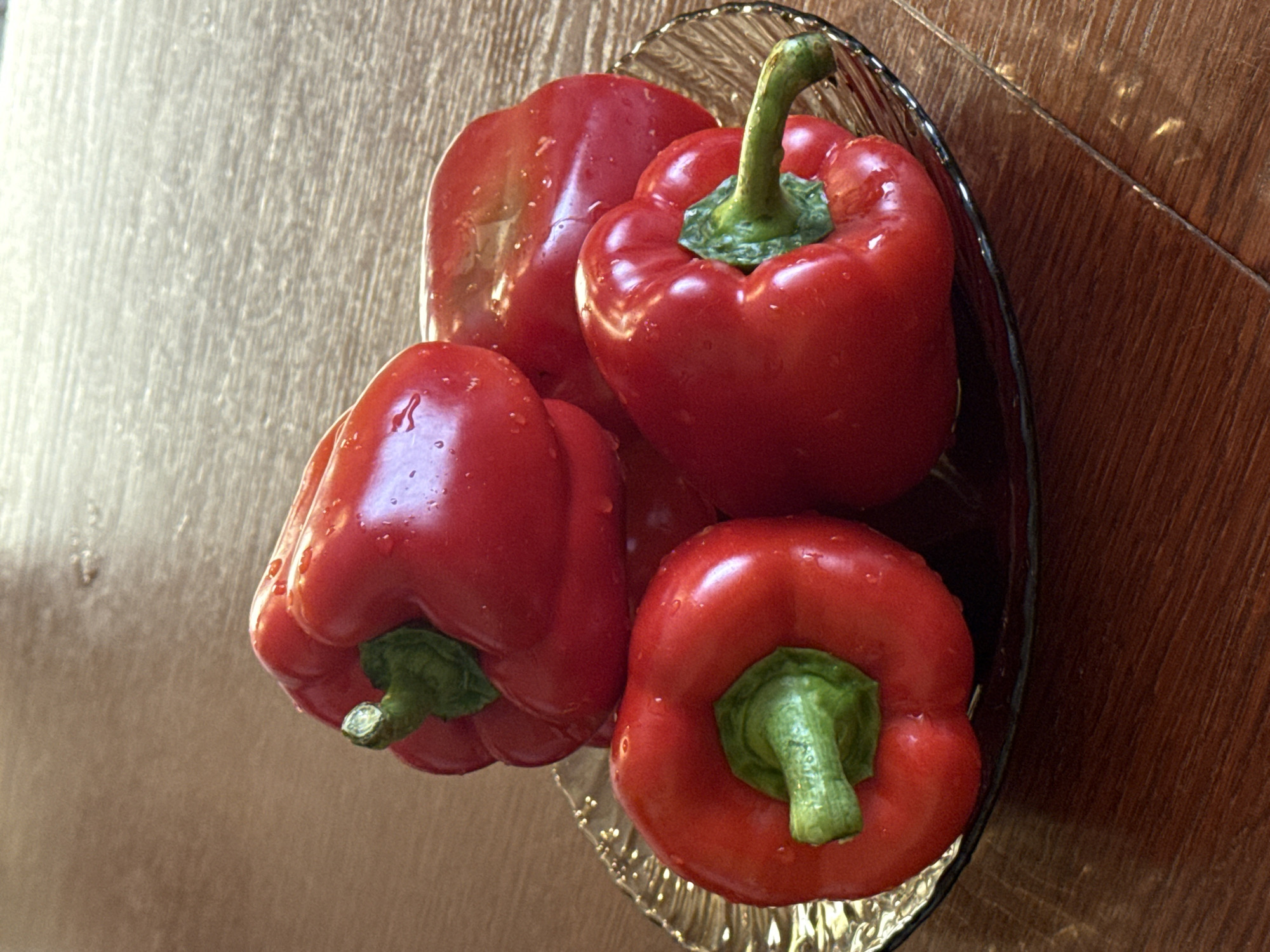 This is a top-down shot of four red bell peppers in a clear glass dish, which is placed on a wooden surface. The peppers are fresh and glistening with water droplets, suggesting they have been recently washed. The lighting appears to be natural, casting subtle shadows and highlighting the textures of the peppers and the wood grain. The scene is simple and domestic, likely from a kitchen or dining area. There is no text visible in the image.