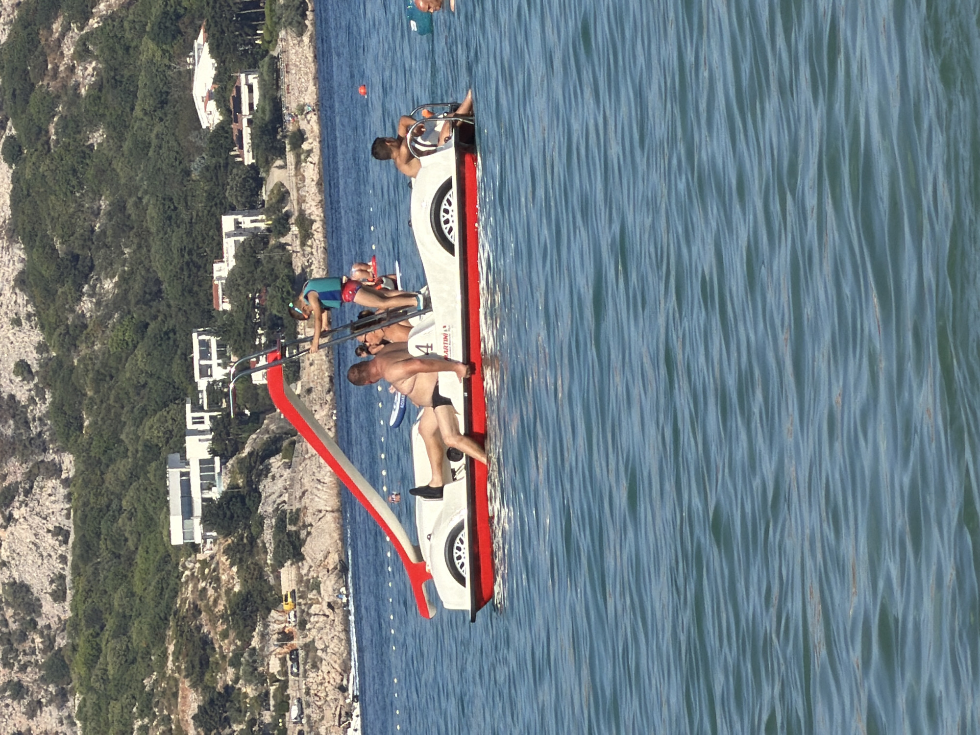 The image captures a pedalo boat with a red and white slide on a clear blue body of water, likely a sea or large lake, under a bright, sunny sky. In the background, a hilly, tree-covered landscape is visible, dotted with modern white buildings and some older structures, indicative of a coastal town or resort.
On the pedalo, there are at least four individuals. A man in black swim shorts is seated on the right side of the boat, with his legs extended. To his left, another person, partially obscured, is leaning against the back of the boat. In the front of the boat, a young person wearing a blue and red life vest is holding onto the ladder of the slide. Another person is seated in the front of the boat, facing away from the camera.
The water is calm with gentle ripples. In the distance, other boats and people are visible, suggesting a recreational area. The presence of the pedalo boat with a slide and the coastal setting strongly suggests this scene is located in a popular tourist area. Given the architecture and landscape, it is plausible this is a location within Općina Baška, Croatia, known for its beaches and coastal tourism.