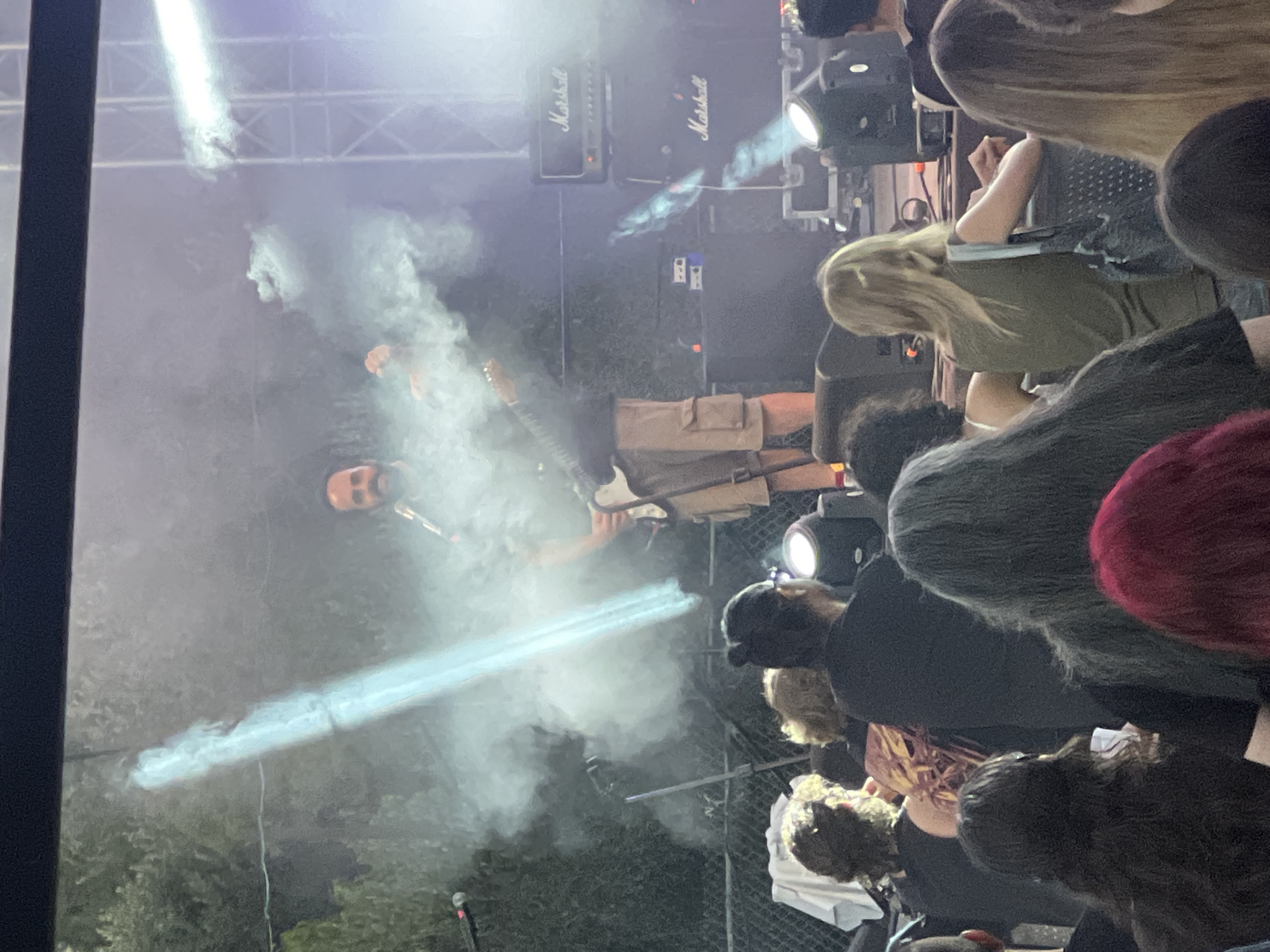 The image depicts a live outdoor music performance at the Analiontas Festival in Analiontas, Cyprus. A male musician, wearing khaki shorts and a dark t-shirt, is on stage playing an electric guitar. He is surrounded by theatrical smoke and illuminated by a bright spotlight. Behind him, two large "Marshall" brand amplifier cabinets are visible. The audience is gathered in front of the stage, with several individuals visible in the foreground, facing the performer. The background includes trees and lighting rigs, indicating an outdoor venue.