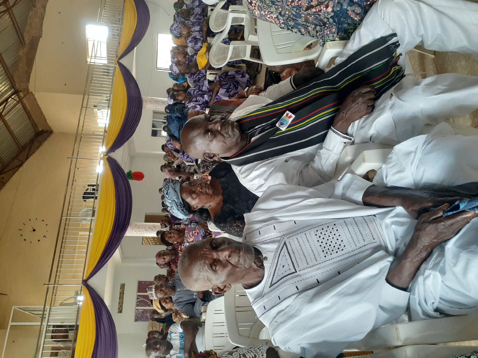 The image shows a gathering of people inside a building, likely in Jos, Nigeria, given the attire of the individuals. Most of the attendees are seated on white plastic chairs. The foreground focuses on two elderly men; one is wearing a white garment with geometric patterns, while the other has a white shirt and a traditional shawl or sash. In the background, a large number of individuals are seated. The room's decor includes yellow and purple draped fabric. A clock is visible on the wall.