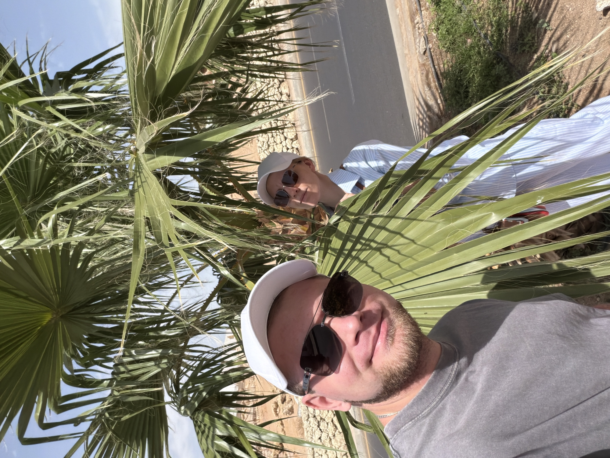 Two people are posing for a selfie amid palm trees. The person in the foreground is a man wearing a white baseball cap, sunglasses, and a gray t-shirt. He has a beard. Behind him is a woman wearing a white baseball cap, sunglasses, and a blue and white striped shirt. They appear to be standing on the side of a road, next to a concrete wall. Beyond the wall is dry, sandy ground with sparse vegetation. The sky is visible through the palm fronds. Given the presence of palm trees and the style of construction nearby, this scene could be consistent with the location of Sharm El Sheikh, Egypt.