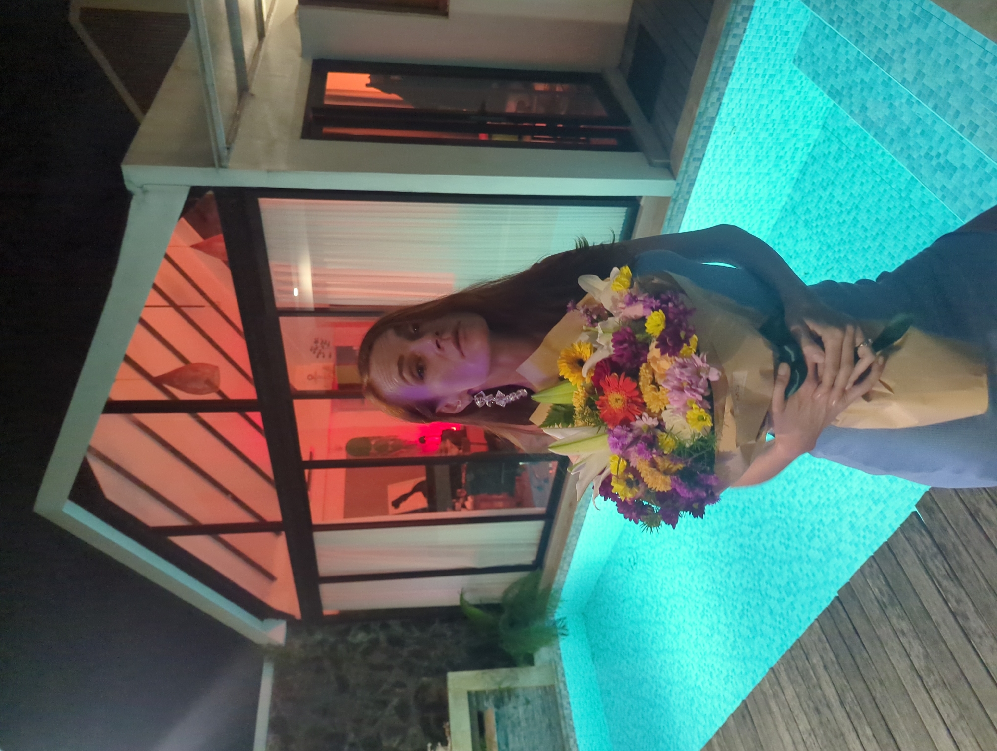 A woman holding a bouquet of flowers stands next to a swimming pool. The background shows a building with orange lighting. The pool is illuminated with a blue-green light. The location is in the city of Kediri, Indonesia.