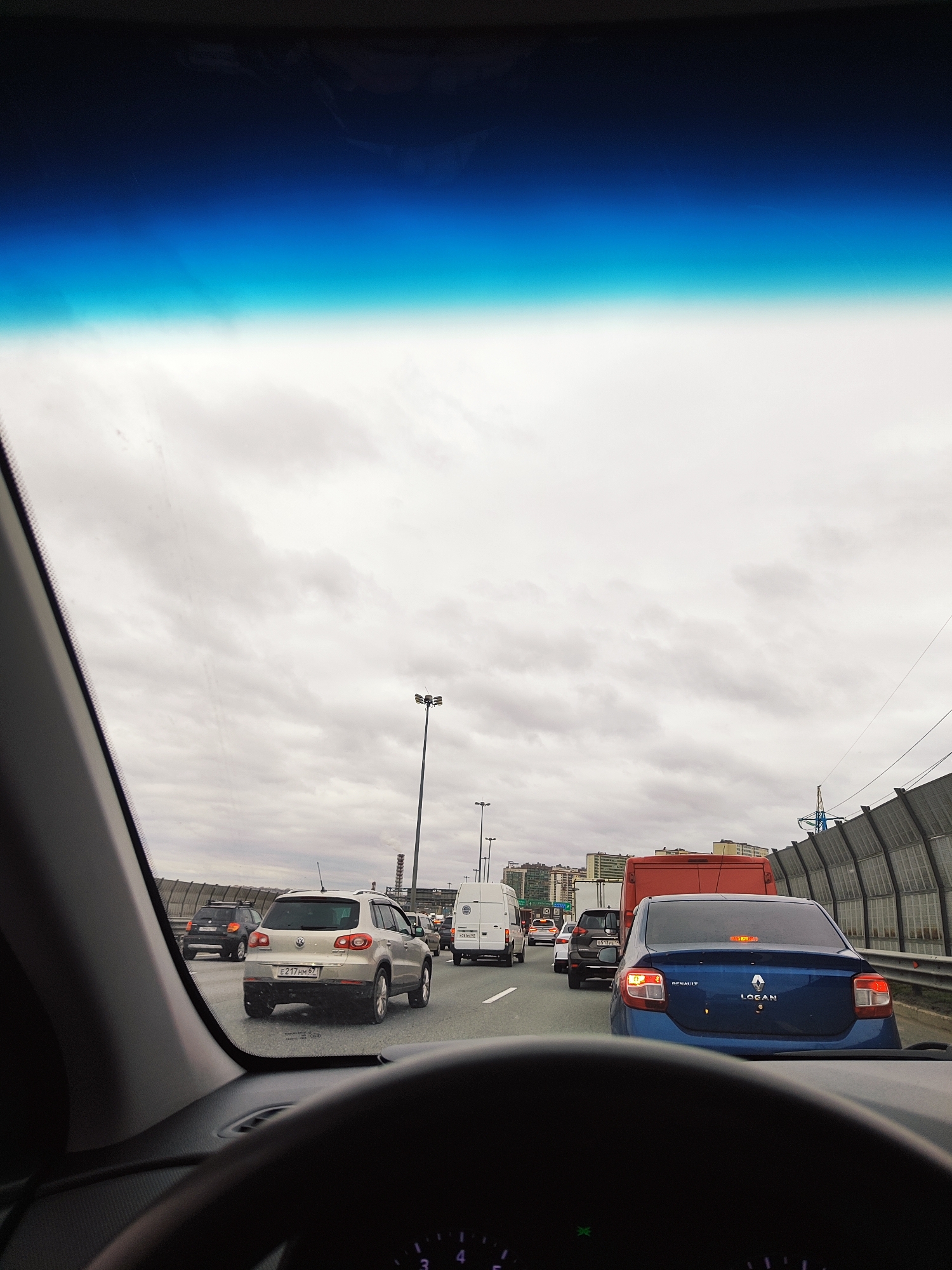 The image provides a driver's perspective from inside a car, looking through the windshield at a multi-lane highway experiencing a traffic jam. The steering wheel and part of the dashboard are visible in the foreground.
Numerous vehicles are present, either stopped or moving slowly. Directly ahead is a silver Volkswagen Tiguan SUV with the Russian license plate `E217HM67`. To its right, a blue Renault Logan sedan is visible, clearly displaying "RENAULT" and "LOGAN" on its trunk. Further ahead in the right lane, a red truck or large van is followed by a white commercial van, which has the Russian license plate `O551YY978`. Other cars are partially visible further down the road in various lanes.
The scene takes place on a wide highway, bordered on the right by tall, grey sound barrier walls. Streetlights with multiple fixtures are spaced along the road. In the distance, several buildings can be seen, hinting at an urban or suburban setting. The sky is heavily overcast and cloudy, indicating a grey daytime, though the specific time of day is not discernible. The presence of Russian license plates, particularly the '978' code associated with Moscow, suggests the location is in Russia, possibly near its capital.