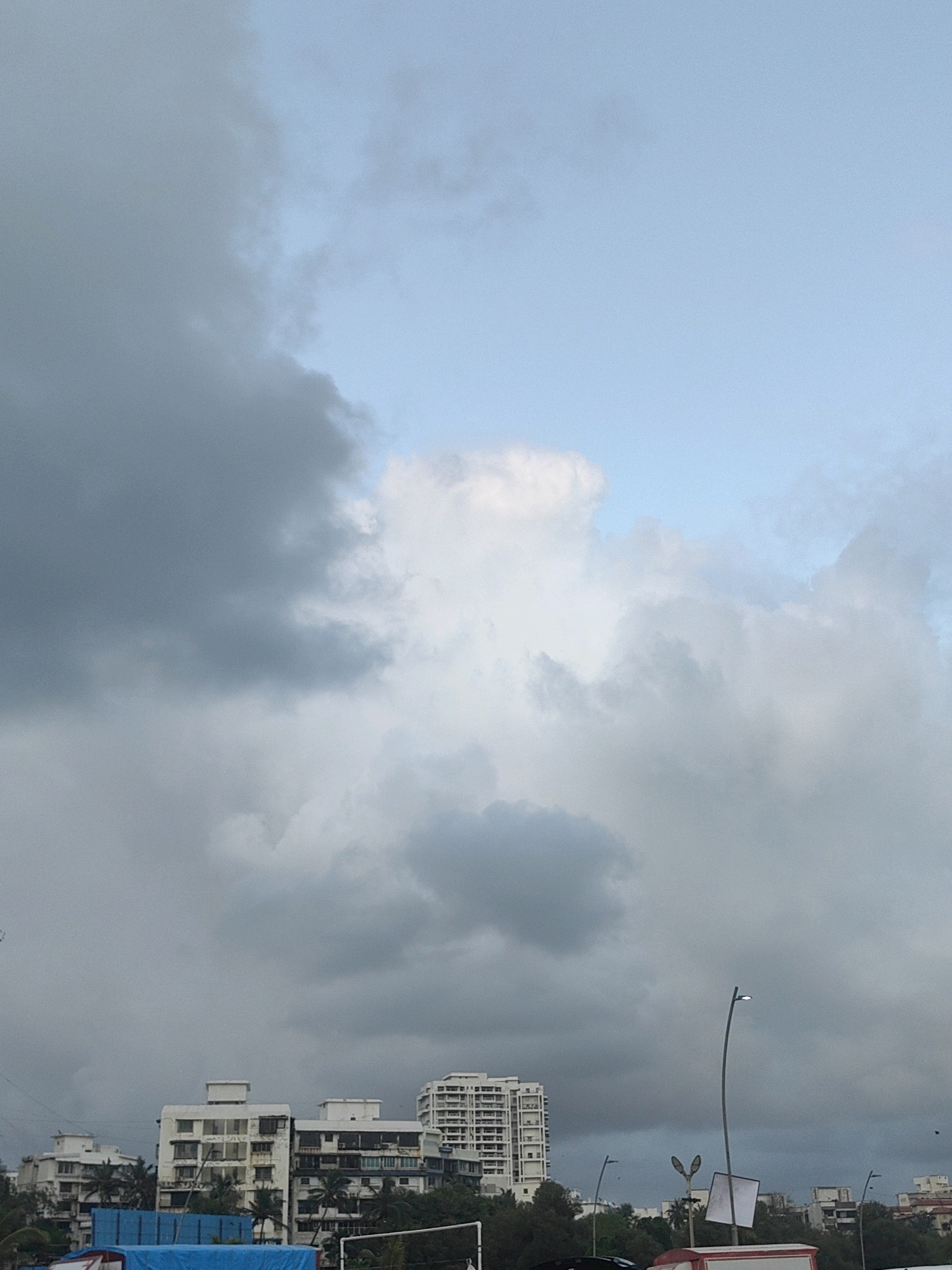 The image captures a cloudy sky over a cityscape, likely in Mumbai, India. In the foreground, the lower half of the image reveals a line of multi-story buildings, some with distinctive architectural features like balconies and window patterns. Trees are visible interspersed between and behind the buildings, some showing the characteristic fronds of palm trees. A bright blue tarp is prominently displayed in the lower left section, suggesting ongoing construction or storage. Streetlights are also visible, with one tall, curved streetlight standing out. In the background, the sky is a mix of heavy, dark gray clouds and lighter, brighter patches, with a sliver of pale blue sky visible at the top. The overall impression is of an overcast day, possibly before or after rainfall.
