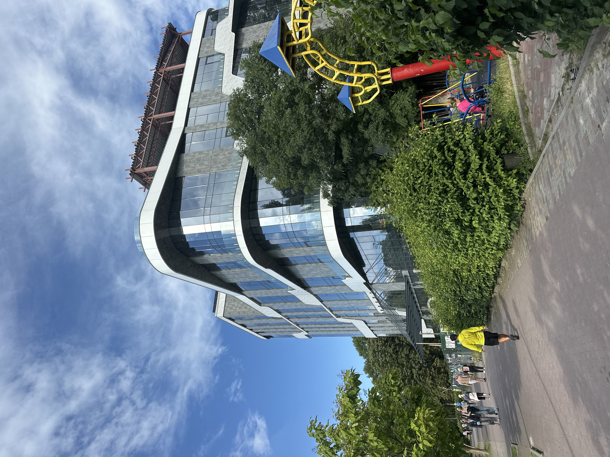 The image depicts a modern, multi-story building with a curved facade, large glass windows, and grey cladding, situated in Ternopil, Ukraine. The building appears to be an office or commercial complex, with a visible structural frame on its rooftop. In the foreground, a paved walkway runs alongside a lush green hedge. To the right of the walkway, a children's playground with yellow and red structures and blue swings is partially visible, with a person in a pink top seated on a swing. Several people are walking on the walkway, including a person in a bright yellow jacket walking away from the camera. The sky is blue with scattered white clouds. The overall scene suggests an urban environment with recreational facilities adjacent to a commercial building.
