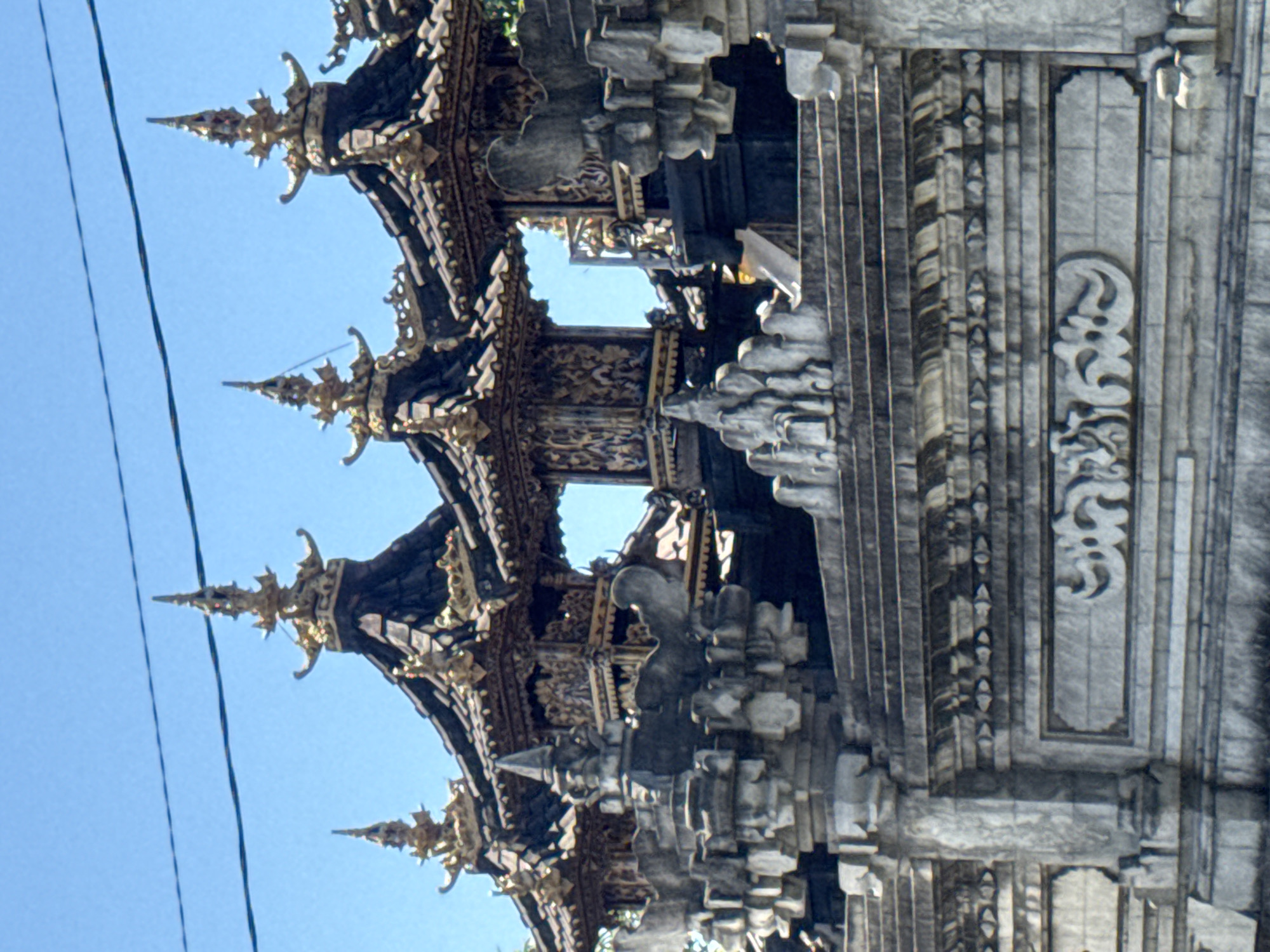 The image depicts a section of an ornate architectural structure in Kubu, Indonesia, likely a temple or ceremonial building. The structure has a multi-tiered roof with intricate carvings and golden accents, set against a clear blue sky. The building is made of grey stone and wood and is richly adorned with sculpted figures and geometric patterns. Powerlines are visible in the top-left corner of the frame.