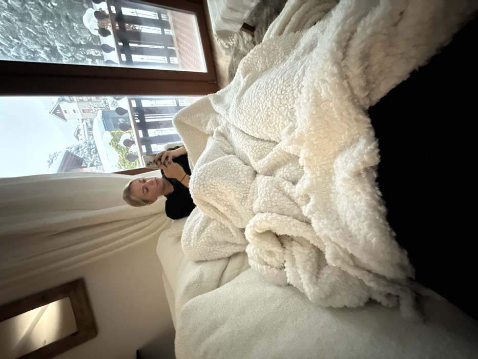A woman lies on a bed near a window, looking at her phone. Snow-covered buildings are visible outside the window. A large, fluffy white blanket dominates the right side of the image, partially obscuring a dark surface. The room has light-colored walls and a wooden-framed mirror is visible in the lower-left corner. Curtains hang near the woman and the window.