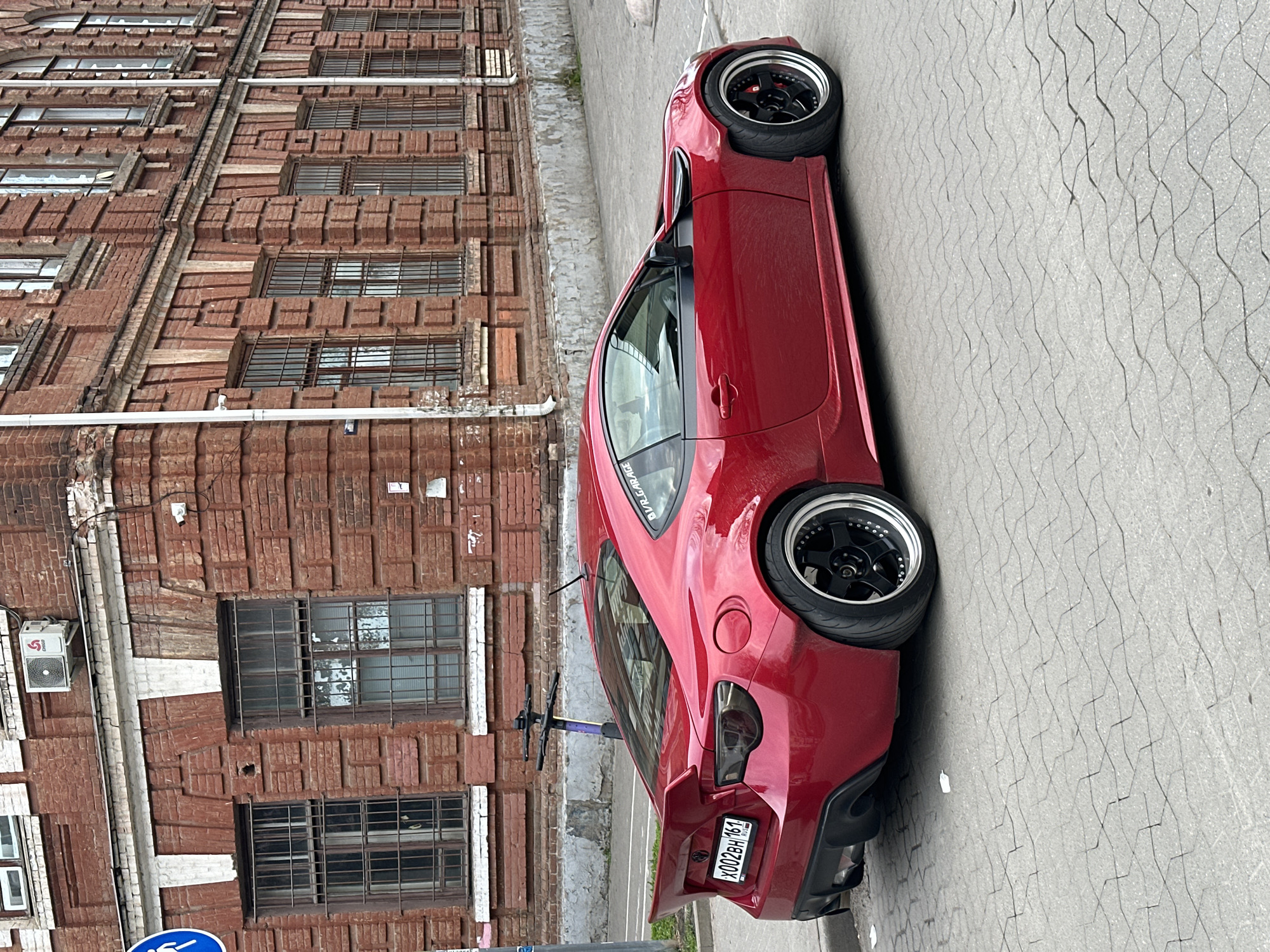 A red sports car is parked on a paved street in Rostov-on-Don, Russia, adjacent to a tall, red brick building with multiple windows. The car is facing towards the right side of the image. The license plate on the car reads "X002BH 161". A bicycle is attached to the car. The building appears to be older and possibly historic, with a visible air conditioning unit attached to the building.