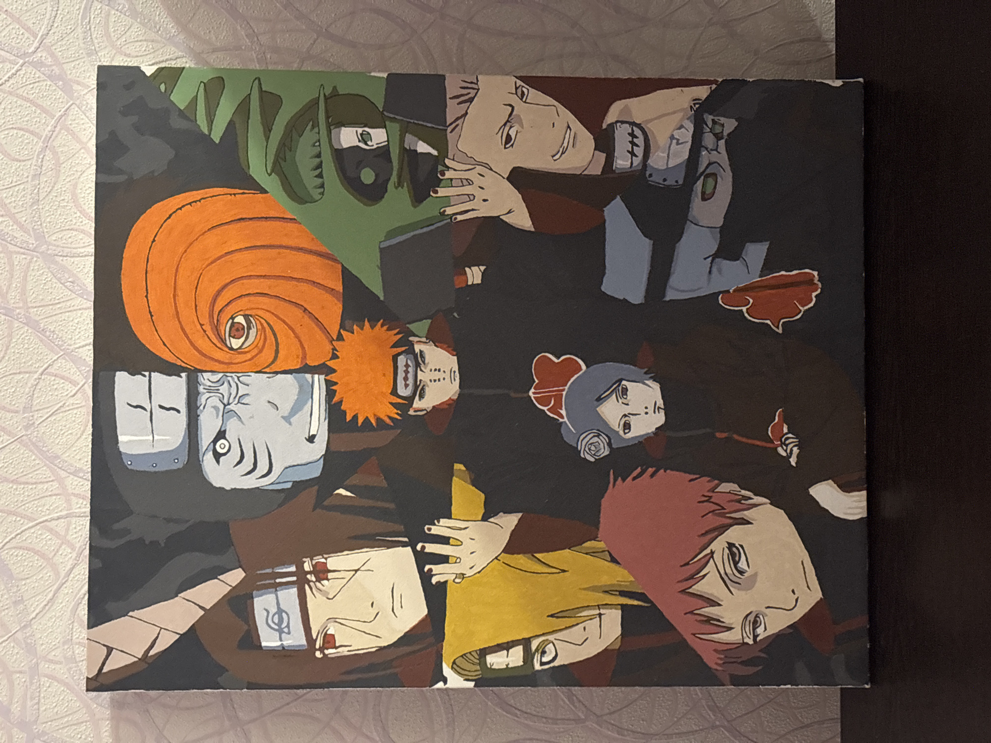 The image shows a fan-made painting of characters from the anime Naruto. The characters, members of the Akatsuki organization, are arranged in a collage-like composition. They include (from left to right, top to bottom):
* Tobi (orange mask)
* Zetsu (green and black face)
* Kisame Hoshigaki (blue face with shark-like features)
* Itachi Uchiha
* Kakuzu
* Pain
* Deidara
* Konan
* Sasori
The background is dark, possibly black. The painting is displayed against a patterned wallpaper on a vertical surface and a dark brown wall on the right side of the picture. The location is Perm, Russia, but the painting itself is the primary subject.