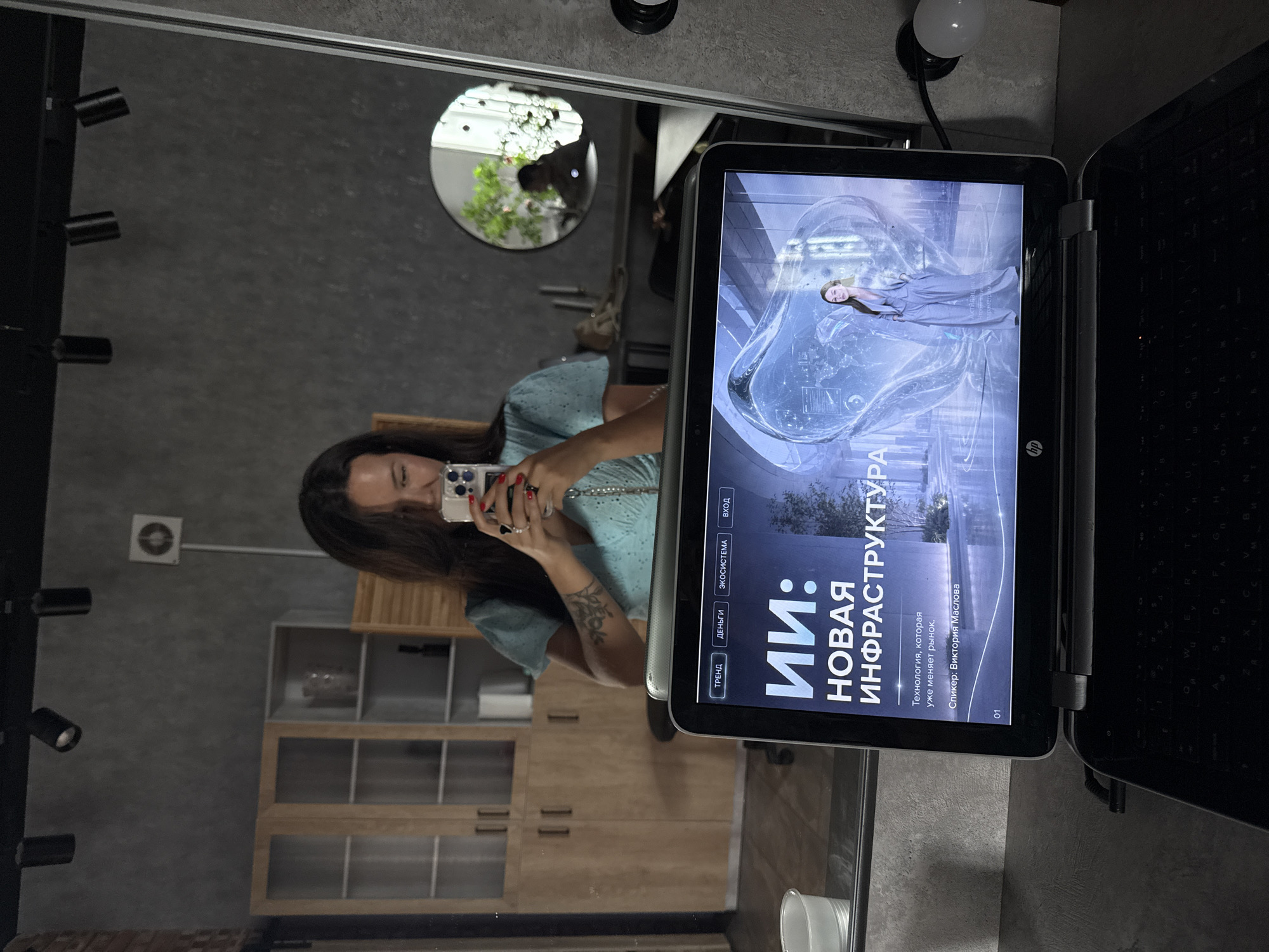 The image depicts a woman taking a selfie in a room with a mirror. She is holding a smartphone and her reflection shows her facing the mirror, with the camera of the phone angled towards her. In the foreground, a laptop is open, displaying a presentation with the title "ИИ: НОВАЯ ИНФРАСТРУКТУРА" which translates to "AI: NEW INFRASTRUCTURE". The presentation also features text that reads "Технология, которая уже меняет рынок" ("Technology that is already changing the market") and "Спикер: Виктория Маслова" ("Speaker: Victoria Maslova").
The environment appears to be an indoor space, possibly an office or studio, with concrete walls and track lighting fixtures on the ceiling. In the background, reflected in the mirror, are wooden cabinets with glass doors, a round mirror showing a potted plant and another person, and some furniture. The overall scene suggests a contemporary, possibly professional or creative setting. There is no discernible information in the image that would allow for an accurate determination of the location within the city of Bely Yar, Russia.
