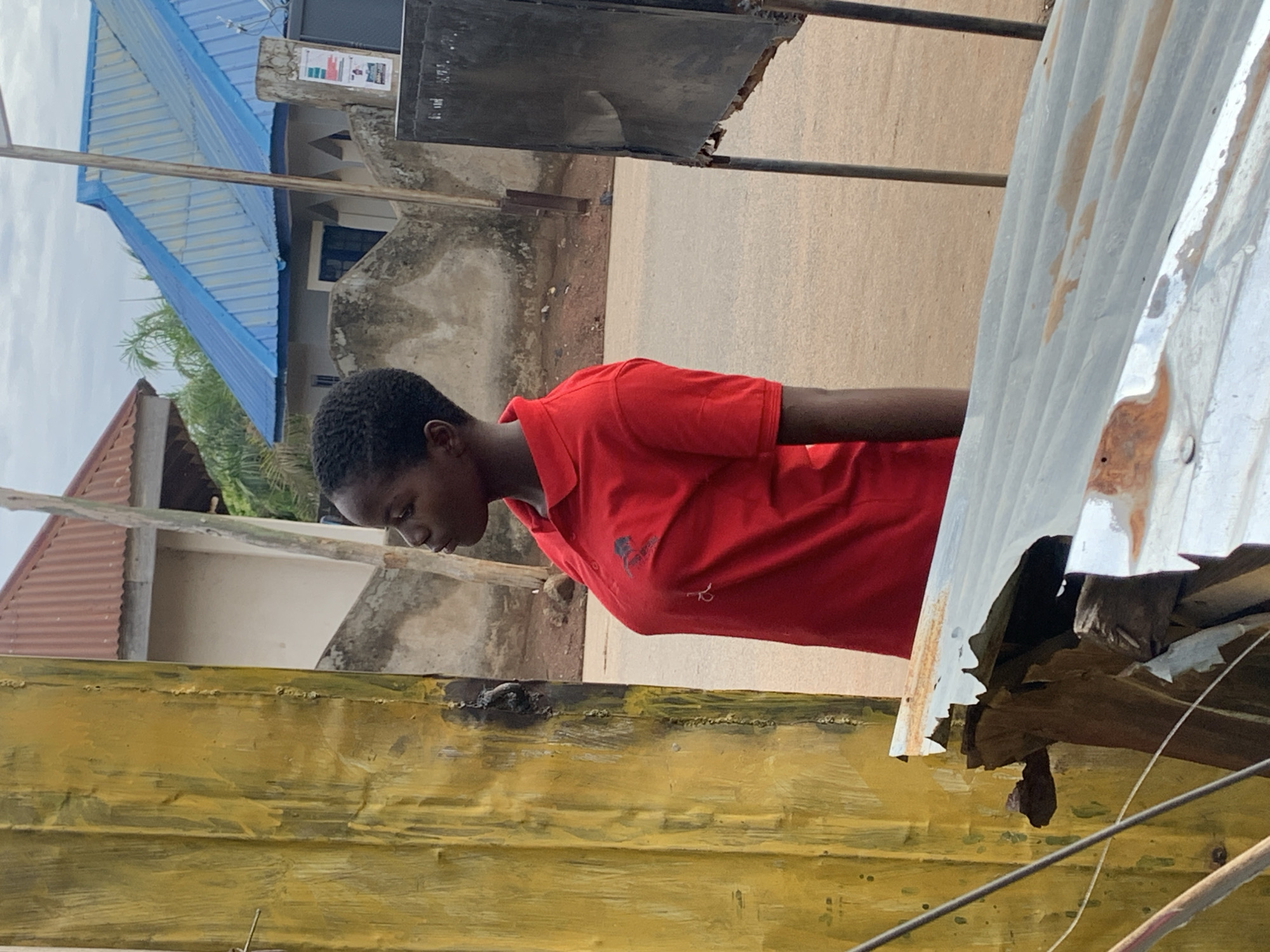 The image shows a person with dark hair and a red polo shirt. The person is outside near a street in what appears to be a residential or commercial area. The background shows buildings with roofs of varying colors. A blue roof is visible towards the top-left of the image. In the foreground, there are elements of what may be constructed structures or materials, including a corrugated metal sheet and yellow painted wood. Based on the prompt, this is likely in Akwanga, Nigeria.